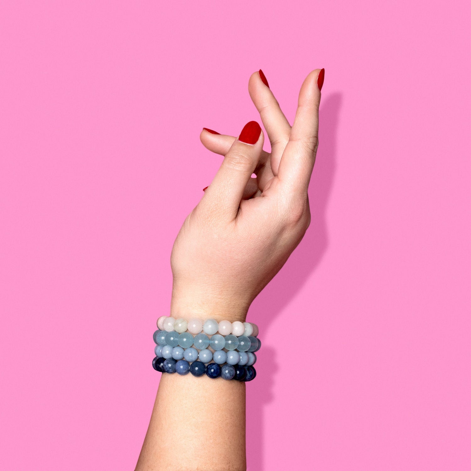 A closeup shot of a stack of blue and pastel crystal gemstone bracelets on a hand with bright red nail polish.