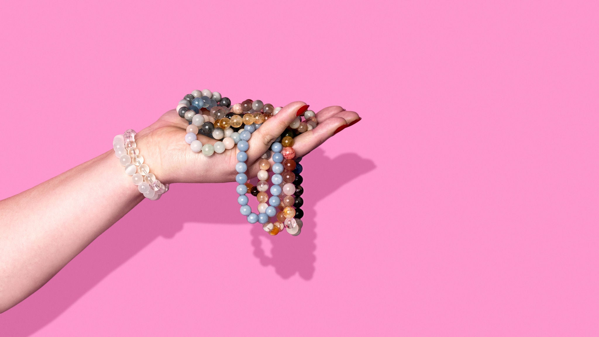 A side profile of a hand on a bright pink background. It wears several crystal bracelets and holds an overflowing pile of colourful gemstone bracelets.