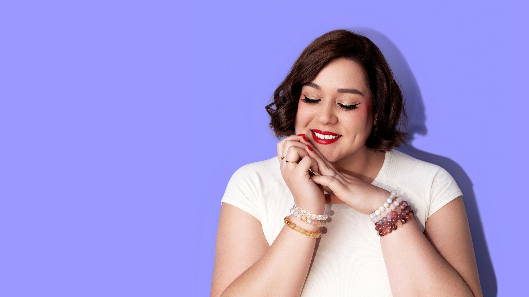 A smiling woman in a white top, with short dark hair and bright red lipstick and nail polish rests her chin on her hands. She wears several gold and white gold rings and a stack of crystal gemstone bracelets on each wrist. The background is bright periwinkle.