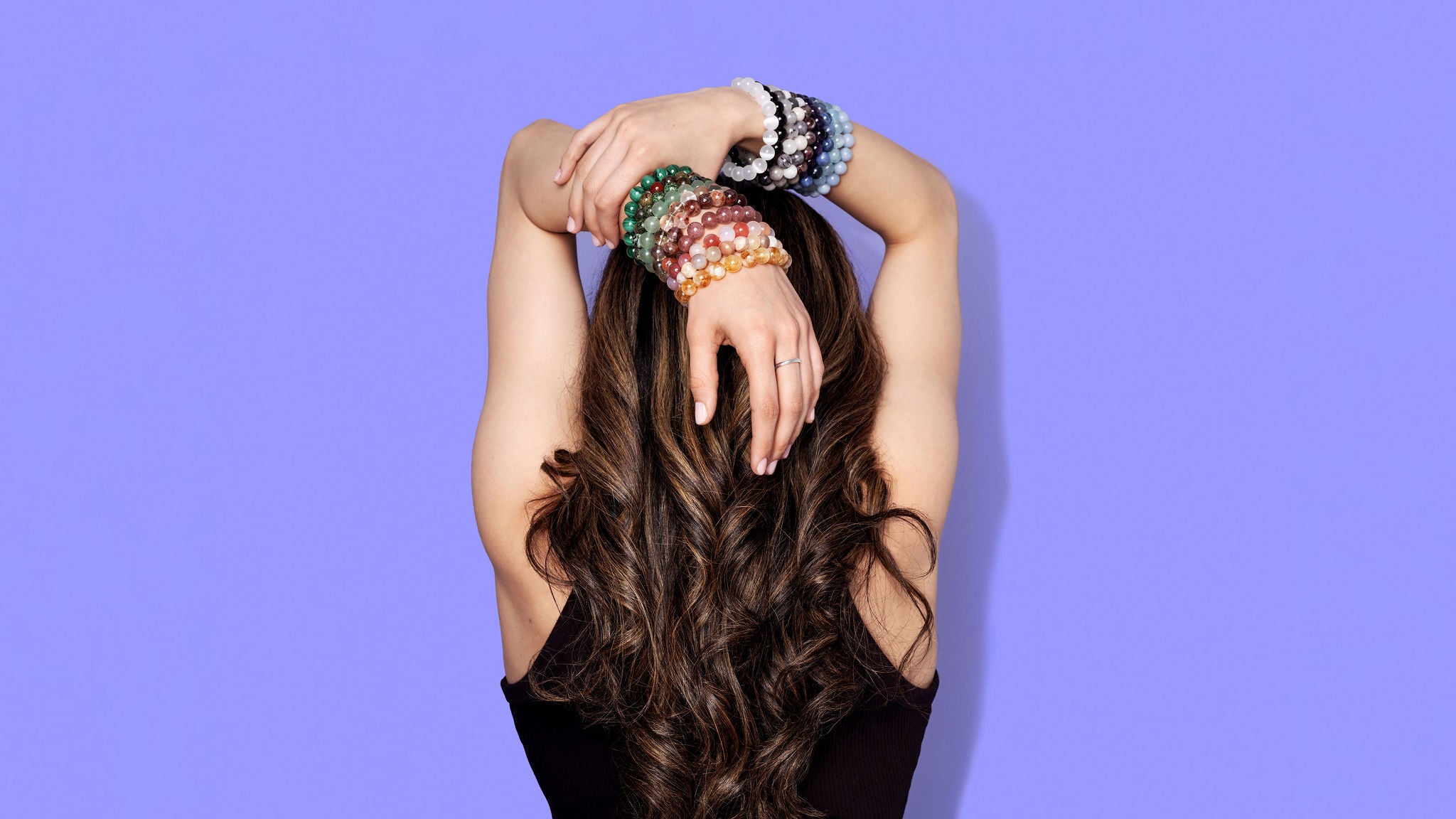 The back of a woman with long dark hair. She has her hands raised over her head with plentiful stacks of colourful crystal gemstone bracelets on each wrist. The background is a bright periwinkle blue.