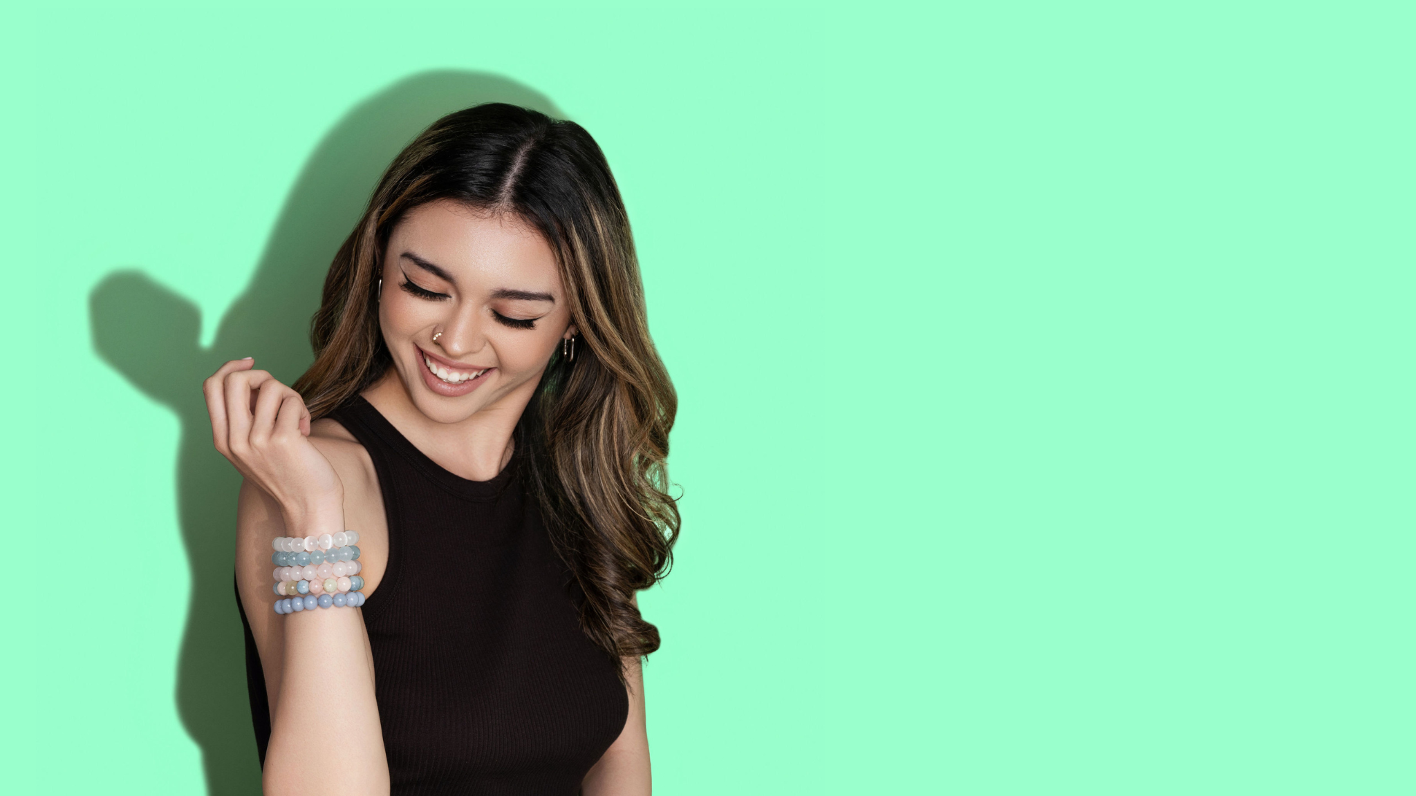 A smiling woman with long hair looks down at a raised wrist. She wears a black top and a large stack of pink, blue, and white crystal gemstone bracelets. The background is bright green.