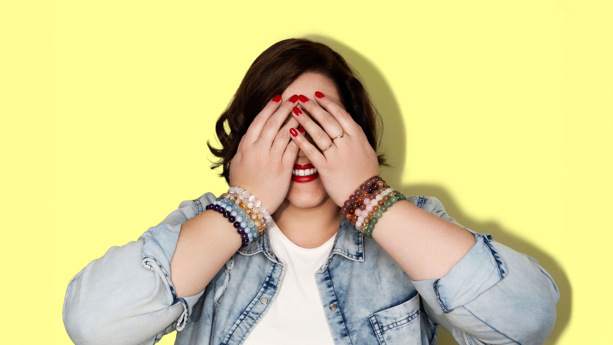 A smiling woman with short dark hair and denim shirt covers her eyes with her hands. She has bright red nail polish and wears two gold rings. On each wrist she has large stacks of colourful gemstone crystal bracelets in red, pink, yellow, green, purple, blue, yellow and white.