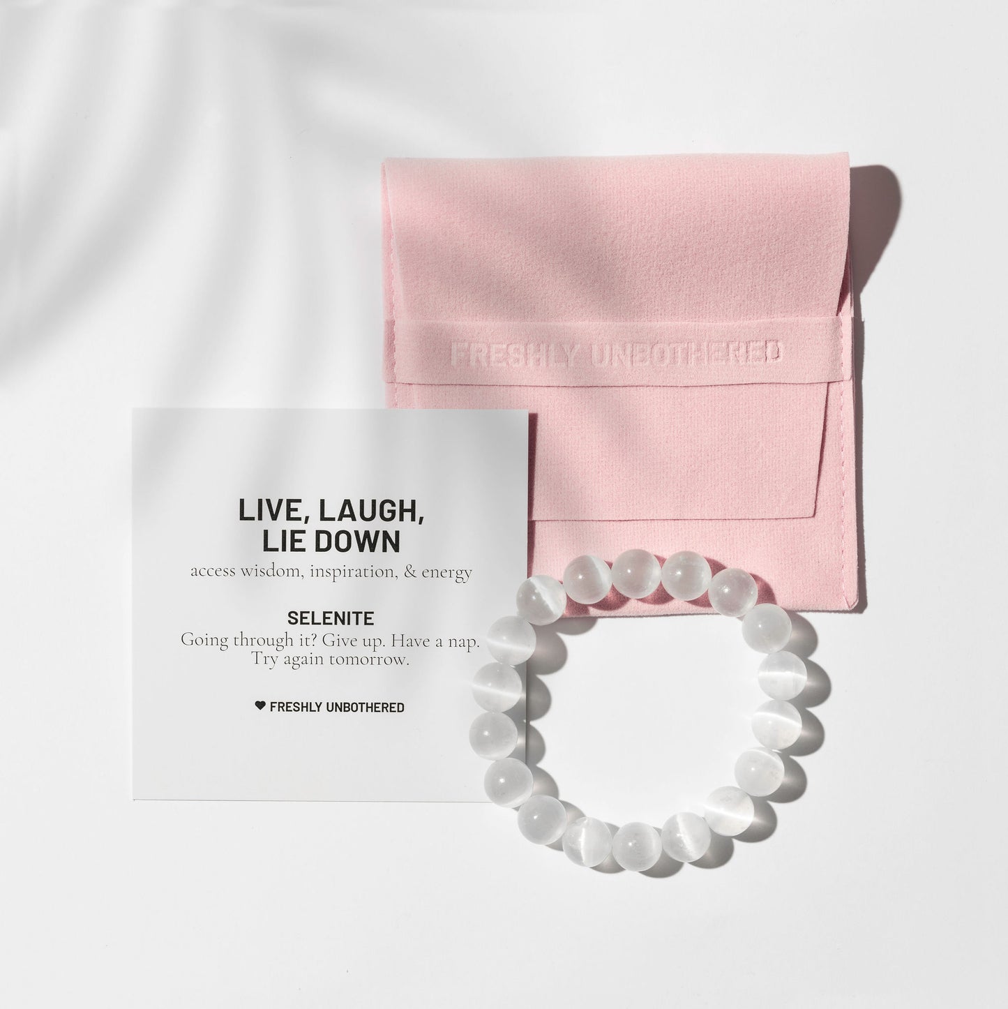 A crystal selenite gemstone bracelet on a white background. There is a square, pink jewellery pouch and a black and white card describing the benefits of the gemstone. There are delicate shadows made by a palm frond.