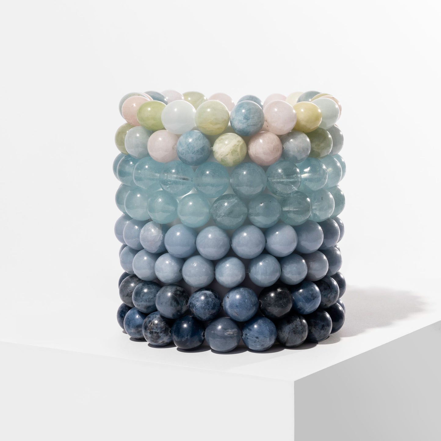 A vertical stack of 8 blue crystal gemstone bracelets, positioned on a white block.