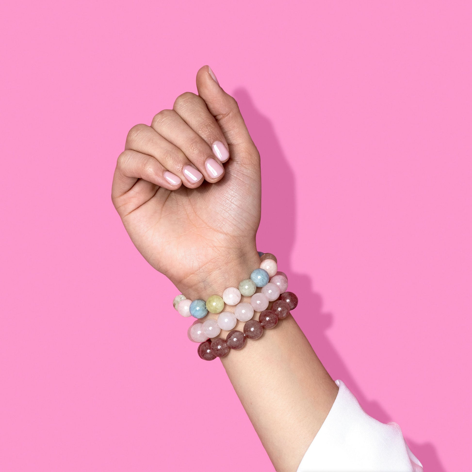 A hand with light pink nail polish wearing 3 different pink shades crystal gemstone bracelets. The background is bright pink with a hard shadow to the right.