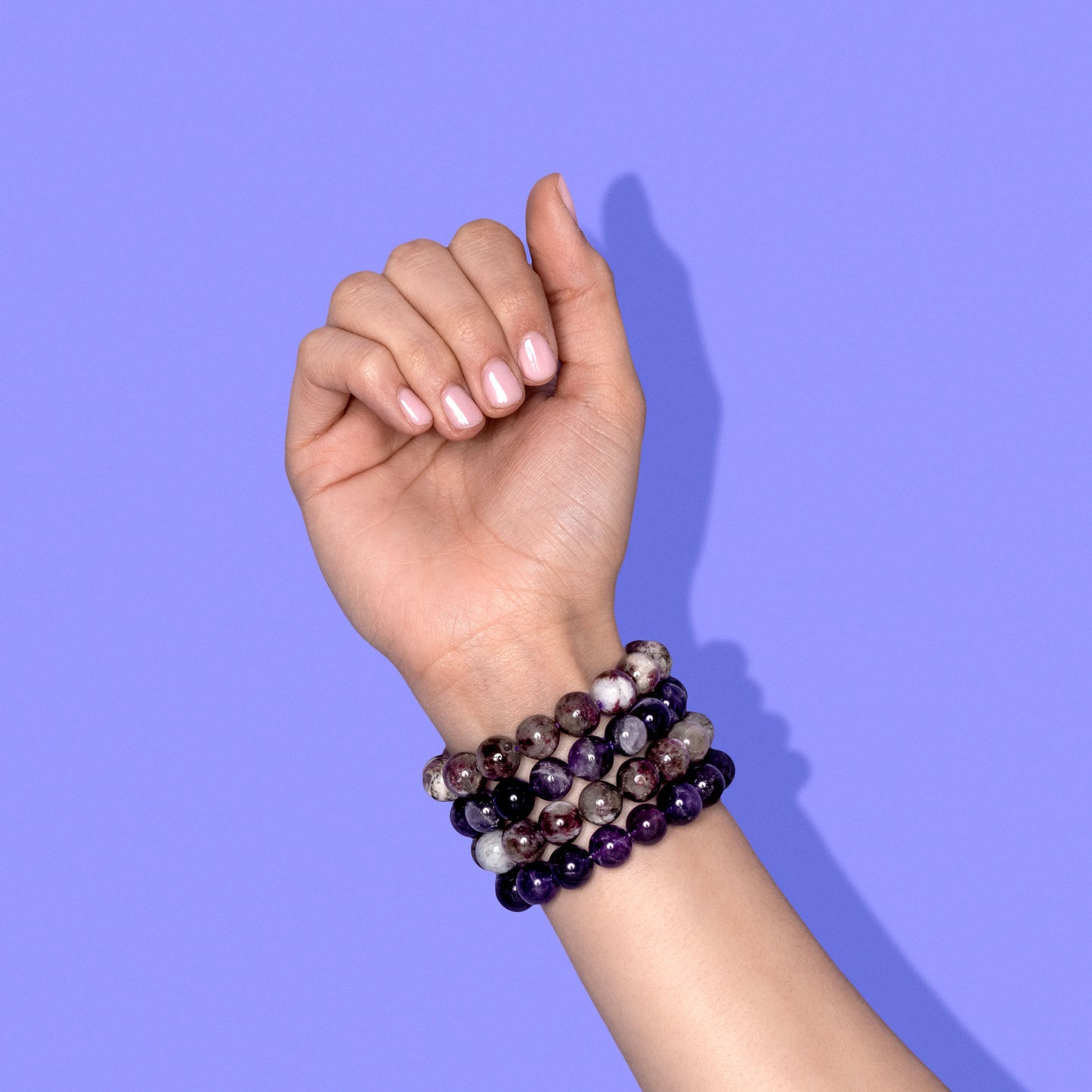 A closeup shot of a stack of purple crystal gemstone bracelets on a hand with light pink nail polish.