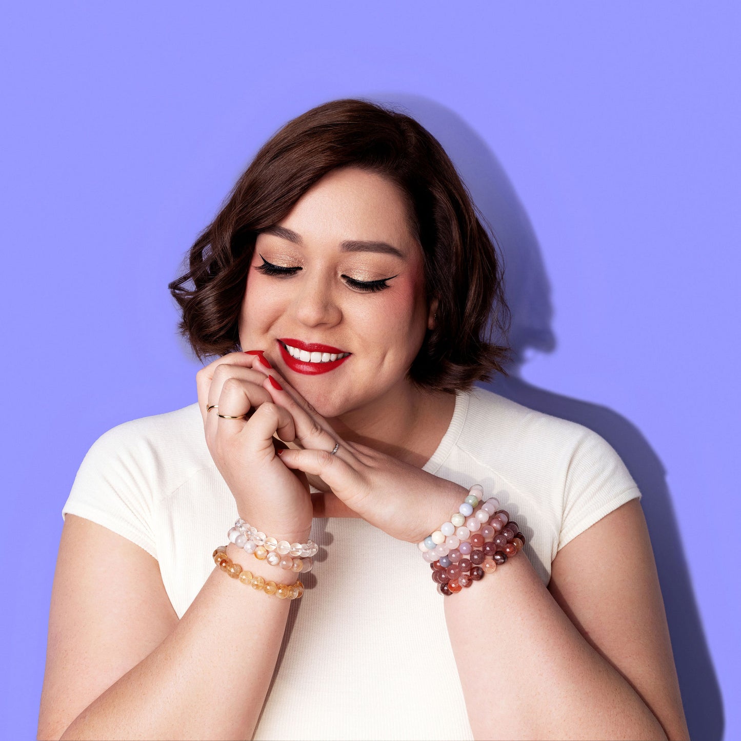 A smiling woman in a white top, with short dark hair and bright red lipstick and nail polish rests her chin on her hands. She wears several gold and white gold rings and a stack of crystal gemstone bracelets on each wrist. The background is bright periwinkle.