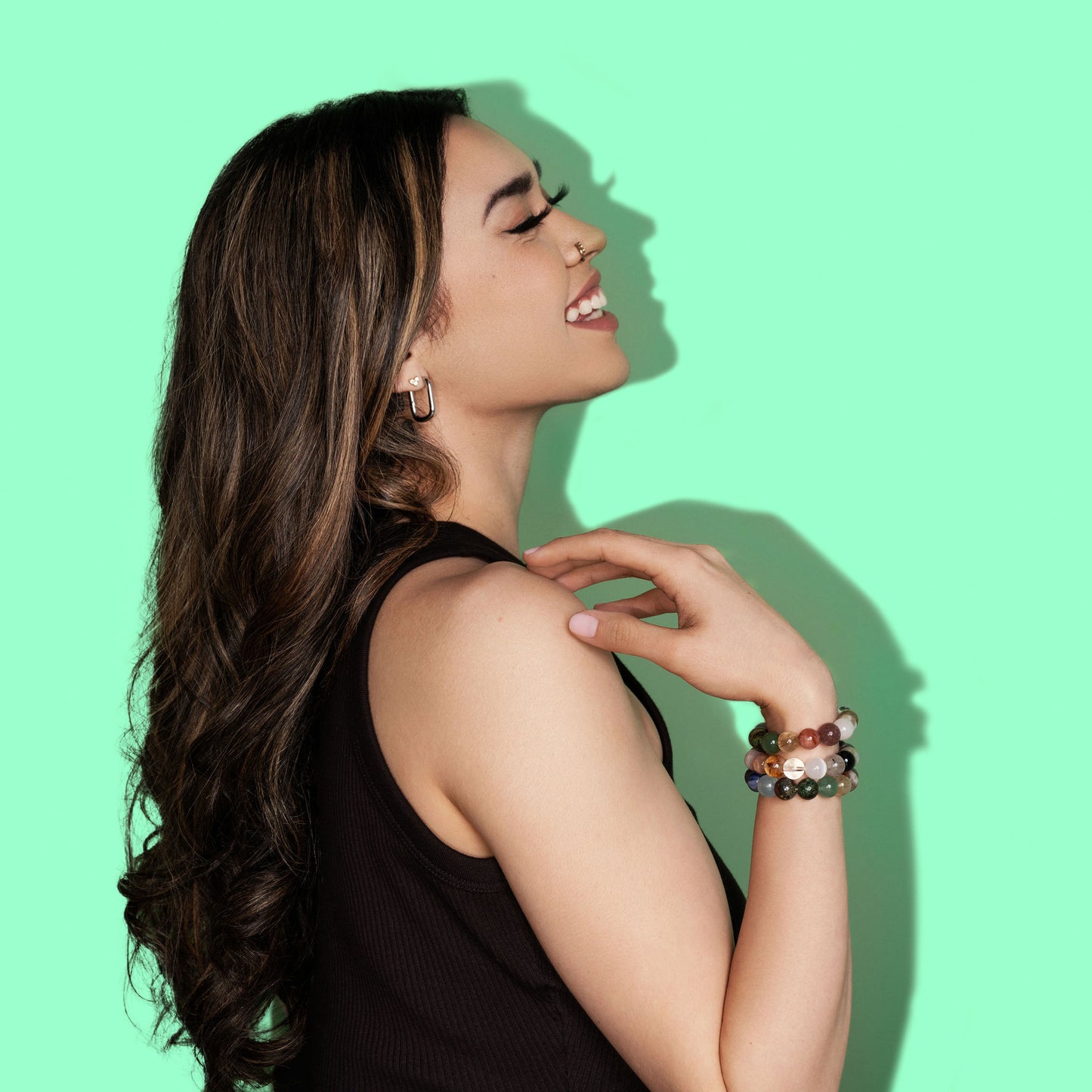 A woman with long dark hair is laughing. She wears a black tank top and is turned to the side and faces to the right. On her wrist she wears several multicoloured crystal gemstone bracelets. The background is a bright green.