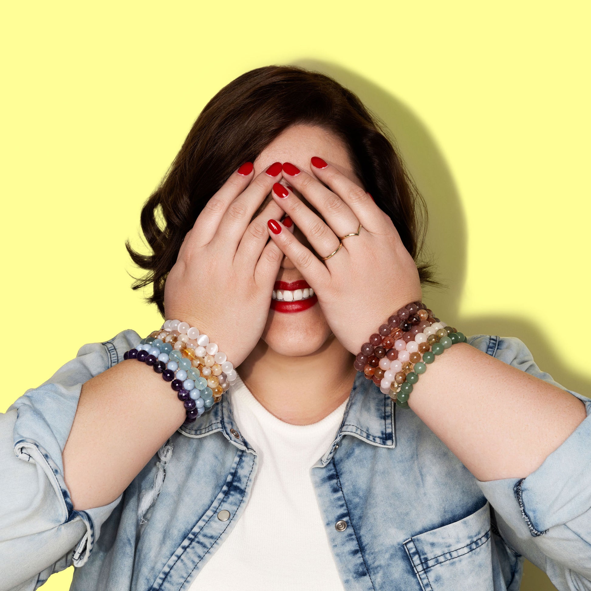 A smiling woman with short dark hair and denim shirt covers her eyes with her hands. She has bright red nail polish and wears two gold rings. On each wrist she has large stacks of colourful gemstone crystal bracelets in red, pink, yellow, green, purple, blue, yellow and white.