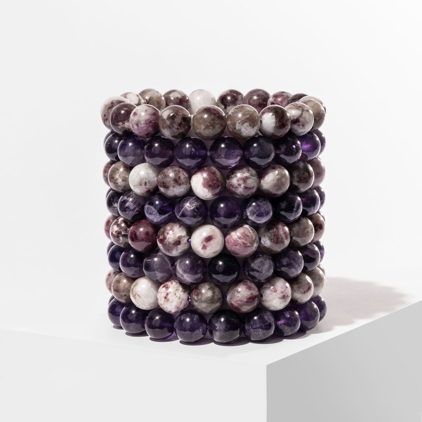 A vertical stack of 8 purple and white crystal gemstone bracelets, positioned on a white block.
