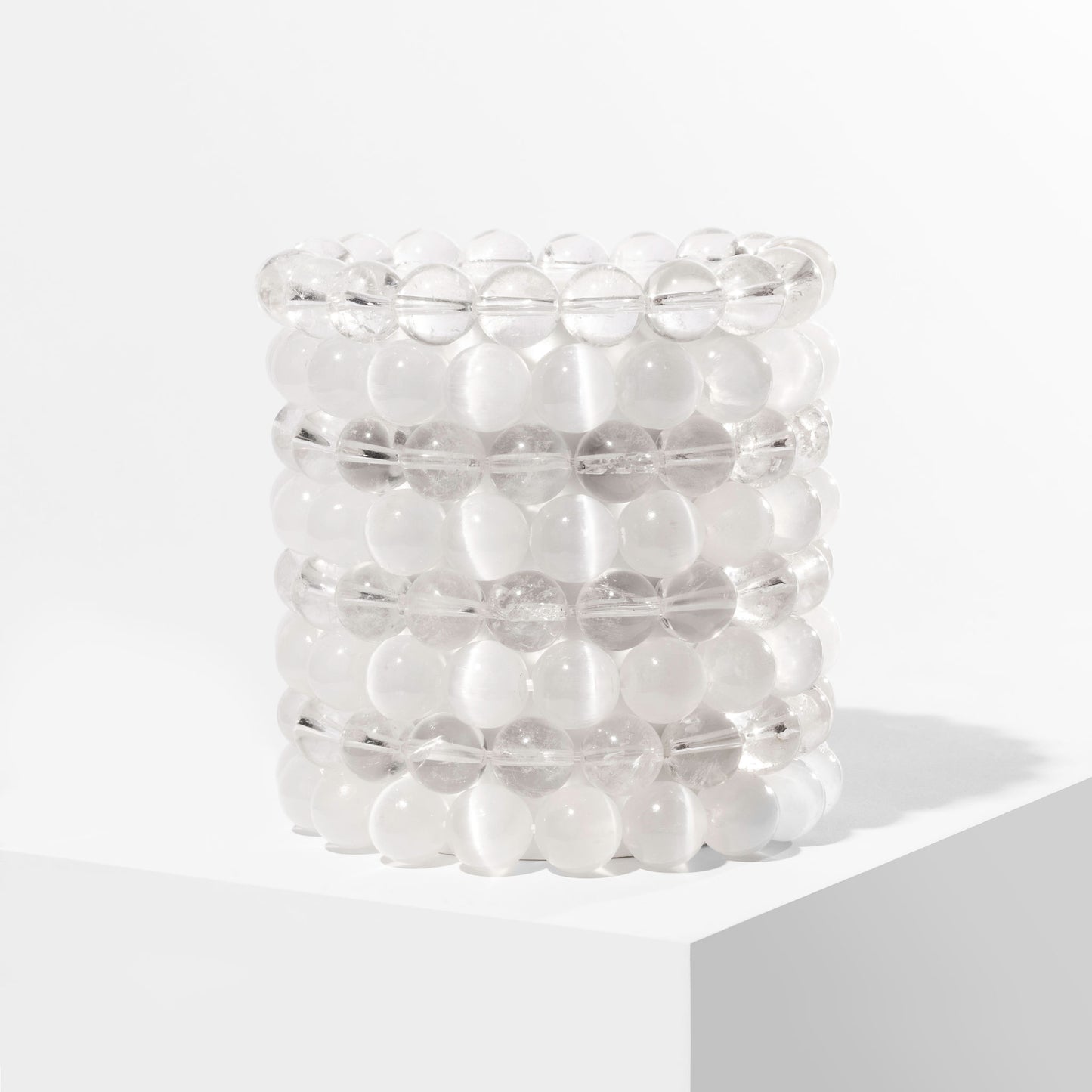 A vertical stack of 8 white and clear crystal gemstone bracelets, positioned on a white block.