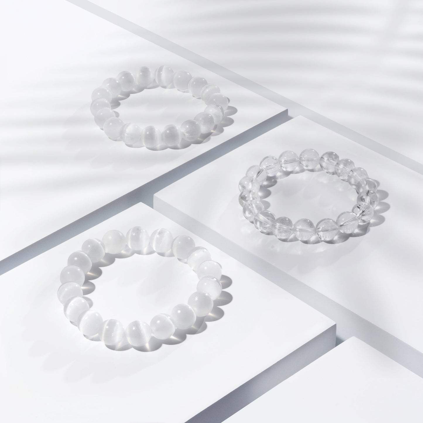 Three white and clear crystal gemstone bracelets positioned on white blocks. There is a palm foliage shadow dappling light across the display.