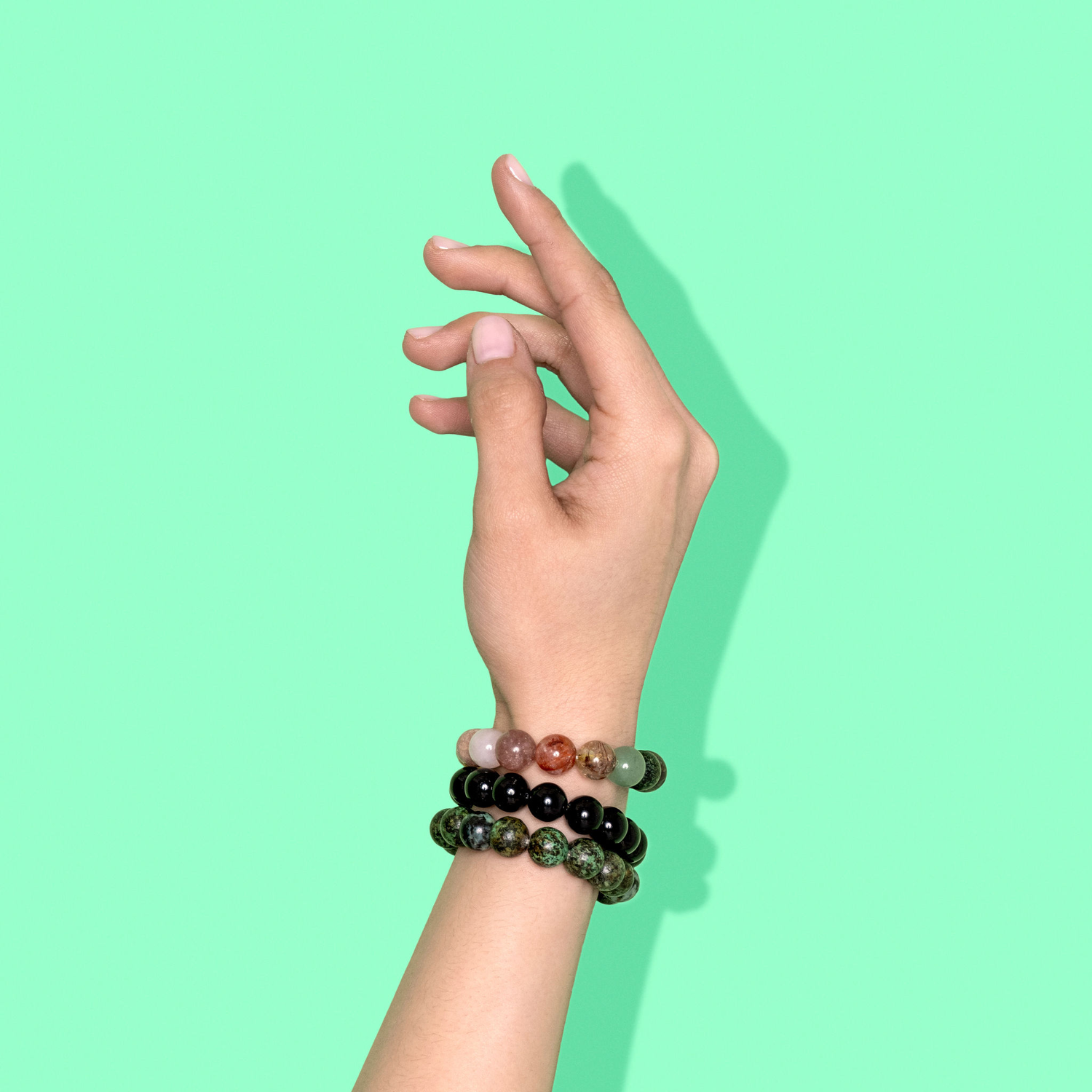 A closeup shot of a stack of green, black, and multicoloured crystal gemstone bracelets on a hand with light pink nail polish.