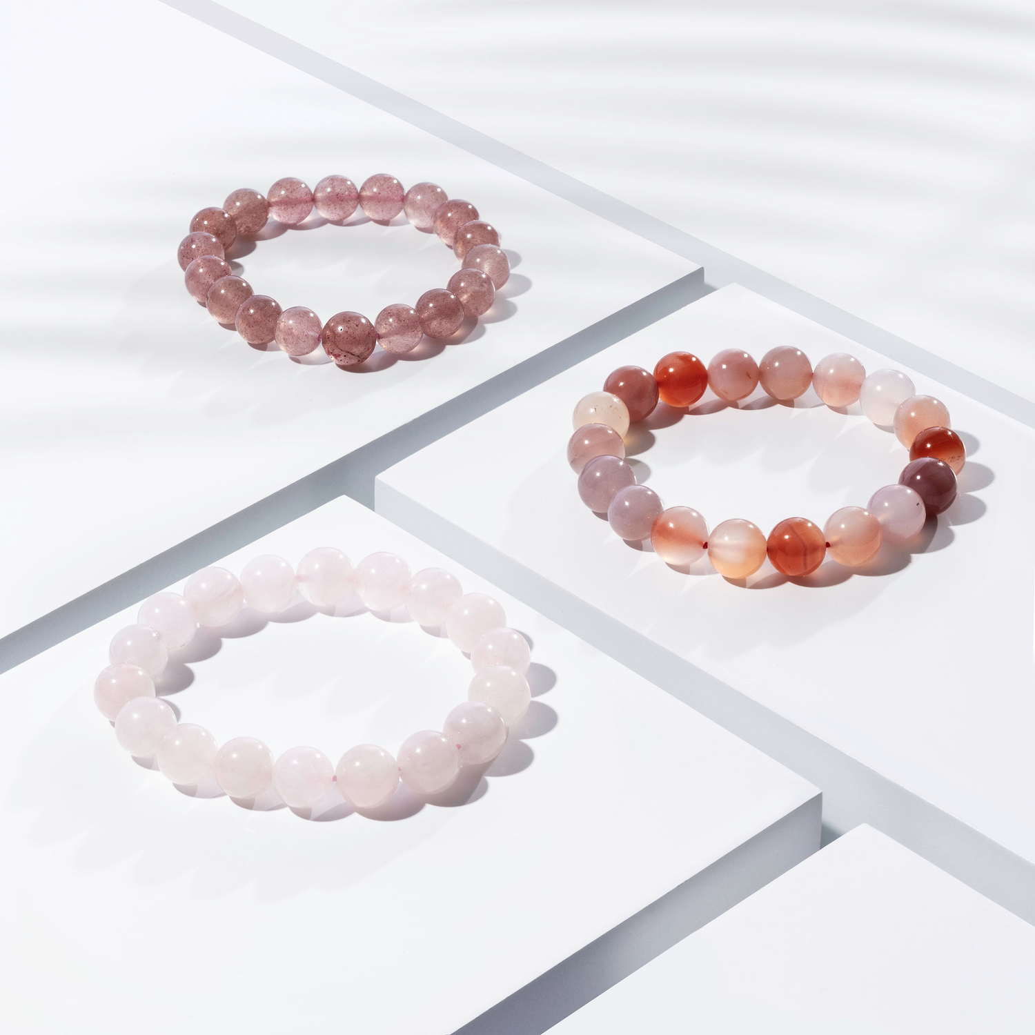 Three various shades of pink coloured crystal gemstone bracelets positioned on white blocks. There is a palm foliage shadow dappling light across the display.