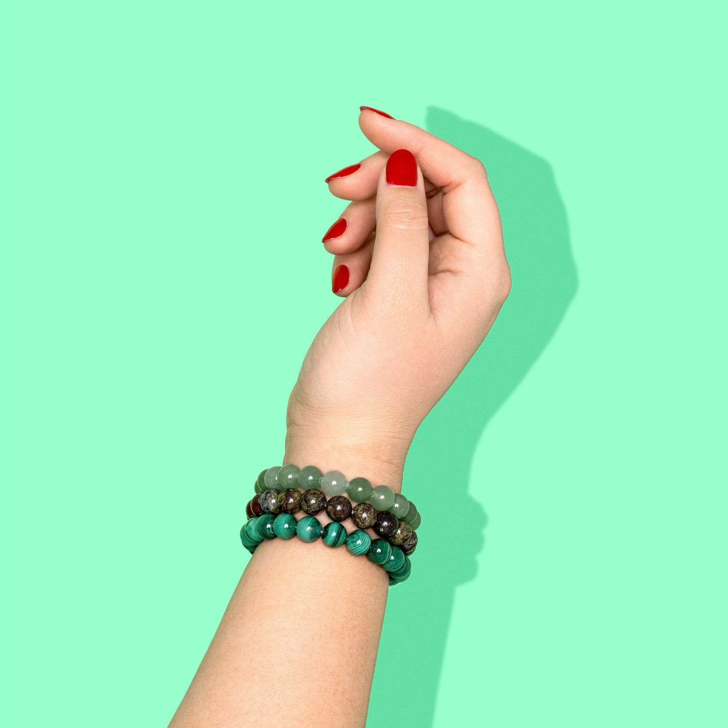 A hand with bright red nails wearing 3 different green coloured crystal gemstone bracelets. The background is bright green with a hard shadow to the right.