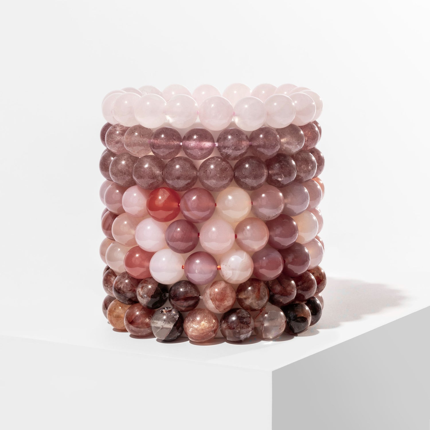 A vertical stack of 8 pink and red coloured crystal gemstone bracelets, positioned on a white block.