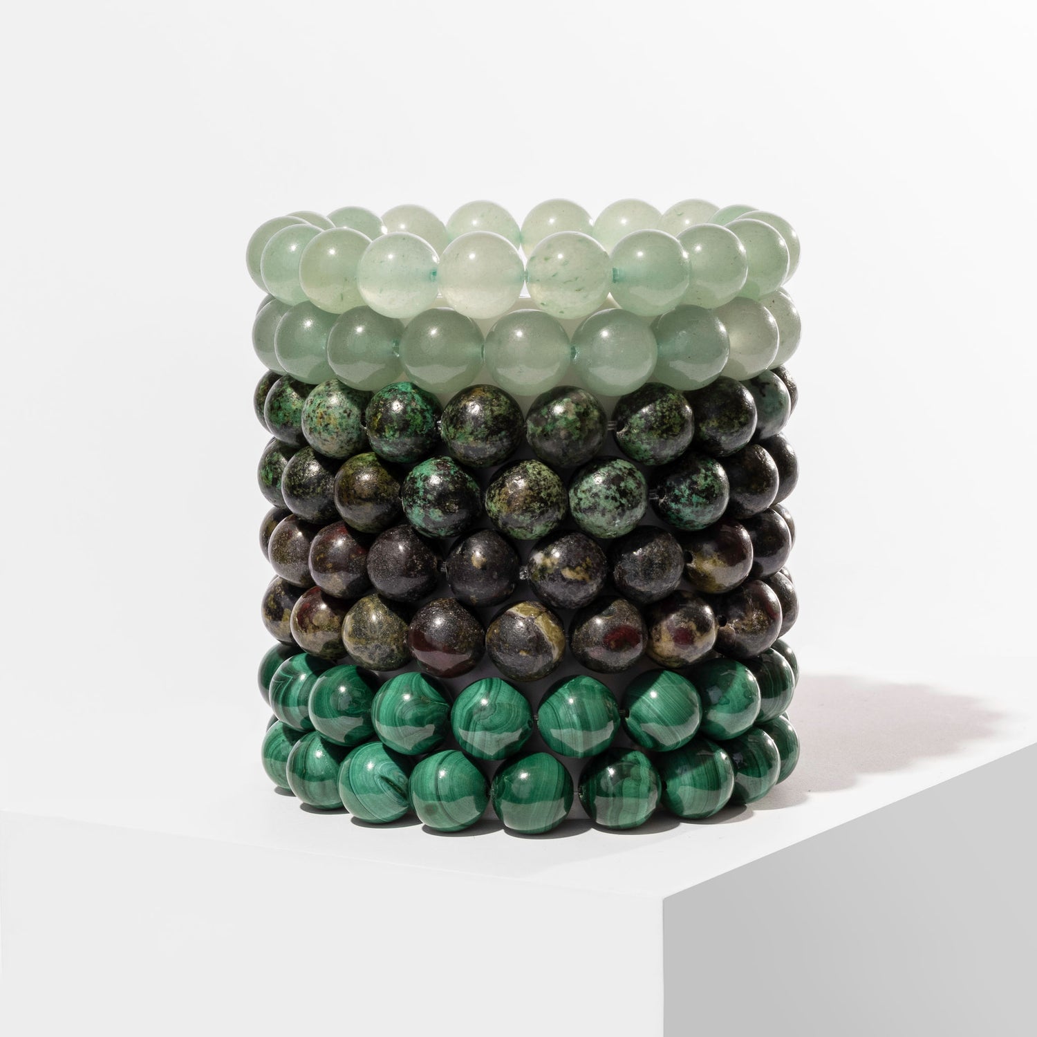 A vertical stack of 8 green coloured crystal gemstone bracelets, positioned on a white block.