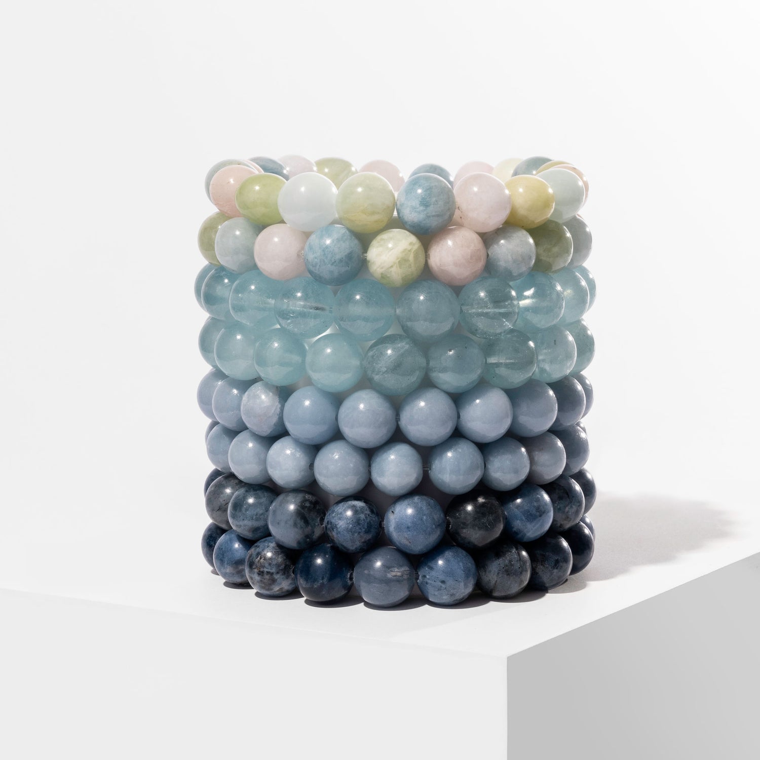 A vertical stack of 8 blue coloured crystal gemstone bracelets, positioned on a white block.