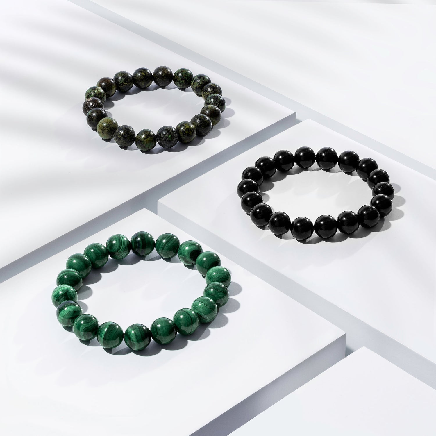 Three green and black coloured crystal gemstone bracelets positioned on white blocks. There is a palm foliage shadow dappling light across the display.