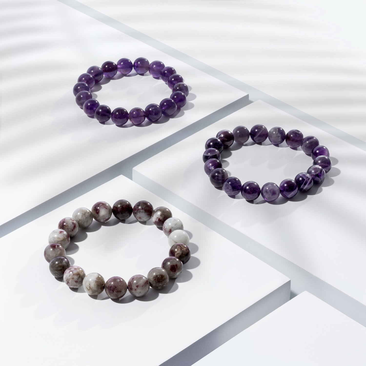 Three purple coloured crystal gemstone bracelets positioned on white blocks. There is a palm foliage shadow dappling light across the display.