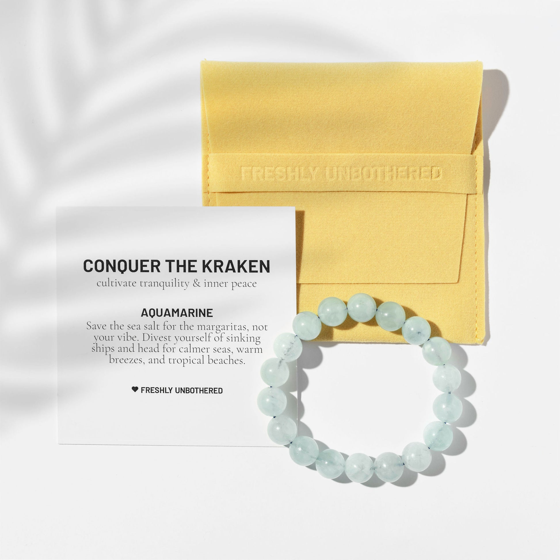 A crystal aquamarine gemstone bracelet on a white background. There is a square, yellow jewellery pouch and a black and white card describing the benefits of the gemstone. There are delicate shadows made by a palm frond.