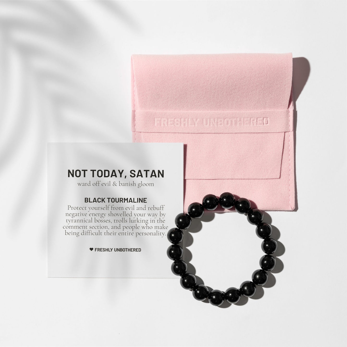 A crystal black tourmaline gemstone bracelet on a white background. There is a square, pink jewellery pouch and a black and white card describing the benefits of the gemstone. There are delicate shadows made by a palm frond.