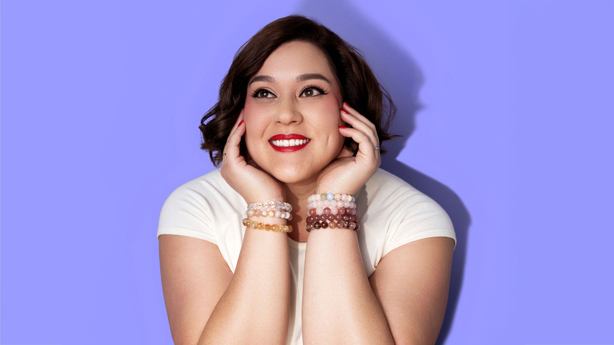 A woman, smiling in bright red lipstick, cups her face with her hands. The background is bright periwinkle. She wears a cream coloured tshirt and on each wrist are numerous crystal gemstone bracelets in a variety of yellow, pink, red, and pastel colours.