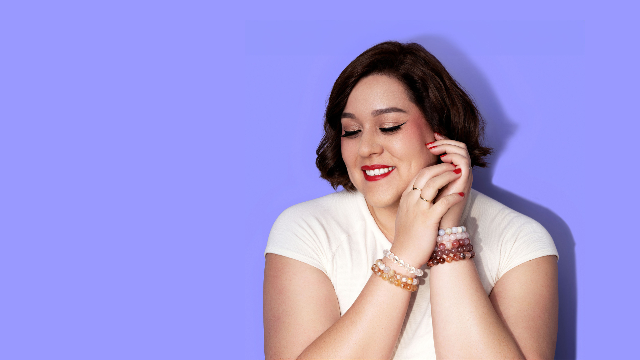 A smiling woman in a white top, with short dark hair and bright red lipstick and nail polish rests her hands to the side of her face. She wears several gold and white gold rings and a stack of crystal gemstone bracelets on each wrist. The background is bright periwinkle.