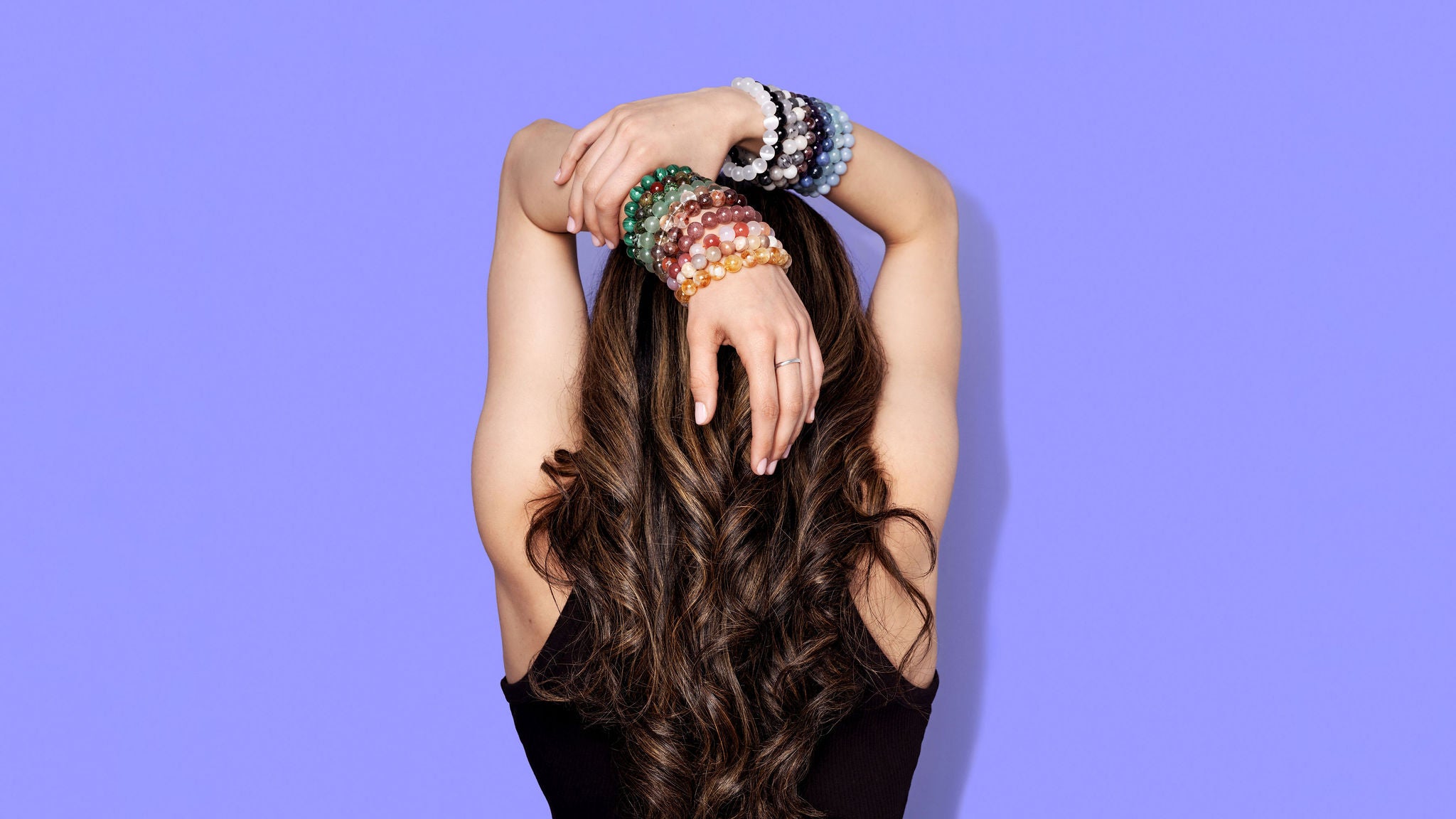 The back of a woman with long dark hair. She has her hands raised over her head with plentiful stacks of colourful crystal gemstone bracelets on each wrist. The background is a bright periwinkle blue. 