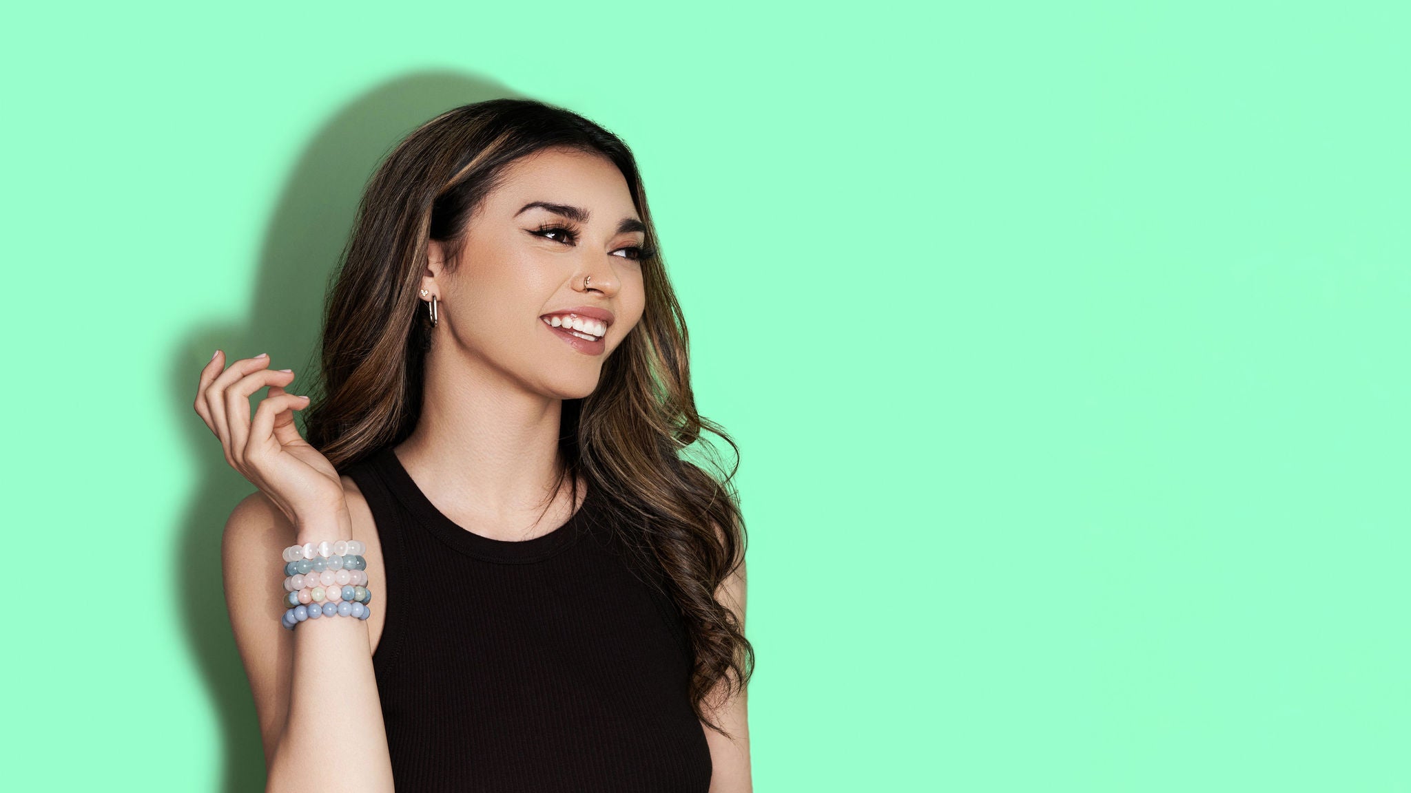 A smiling woman with long dark hair wears a black top and a variety of white, blue, and pink coloured crystal gemstone bracelets. The background is bright green.