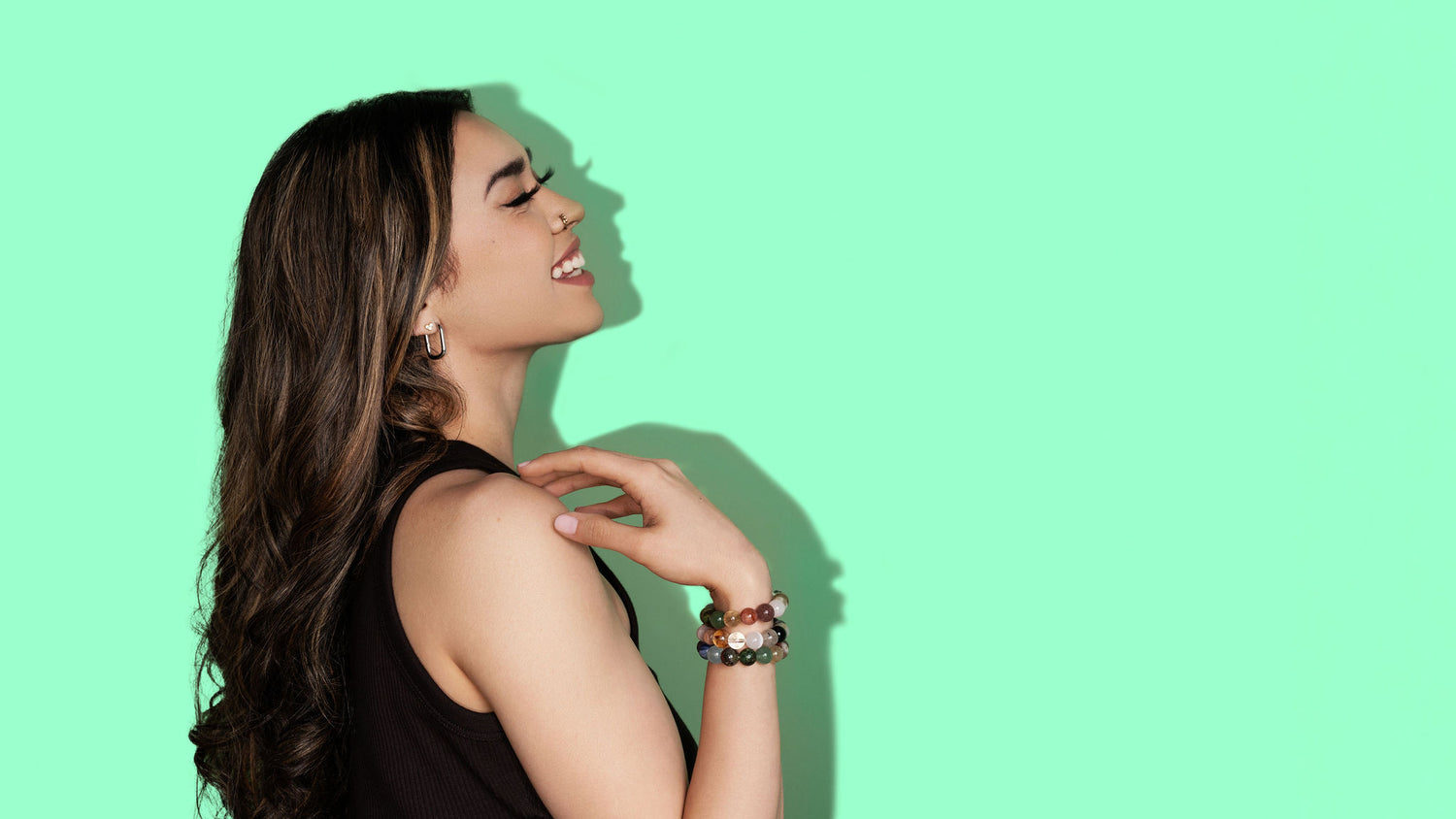 A woman with long dark hair is laughing. She wears a black tank top and is turned to the side and faces to the right. On her wrist she wears several multicoloured crystal gemstone bracelets. The background is a bright green.