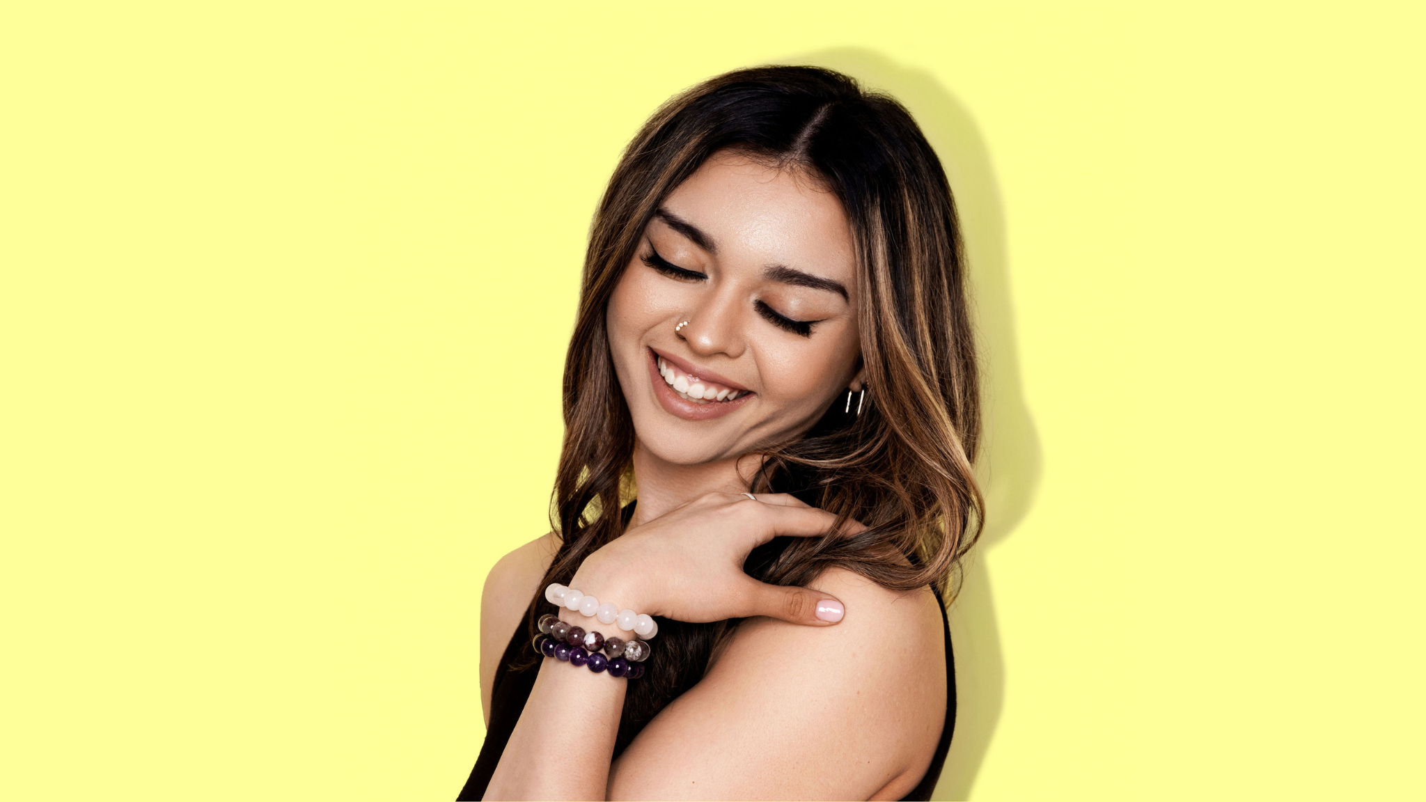 A close up shot of woman with long dark hair wearing a black top, smiles. She has her gaze cast downwards and rests her hand on her shoulder. She is wearing one pink and two purple coloured crystal gemstone bracelets.