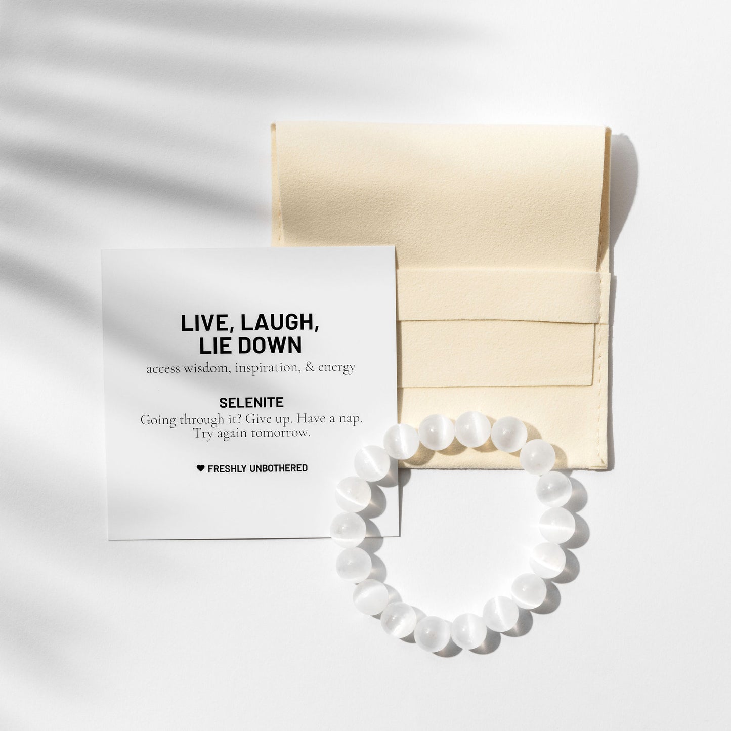 A crystal selenite gemstone bracelet on a white background. There is a square, yellow jewellery pouch and a black and white card describing the benefits of the gemstone. There are delicate shadows made by a palm frond.