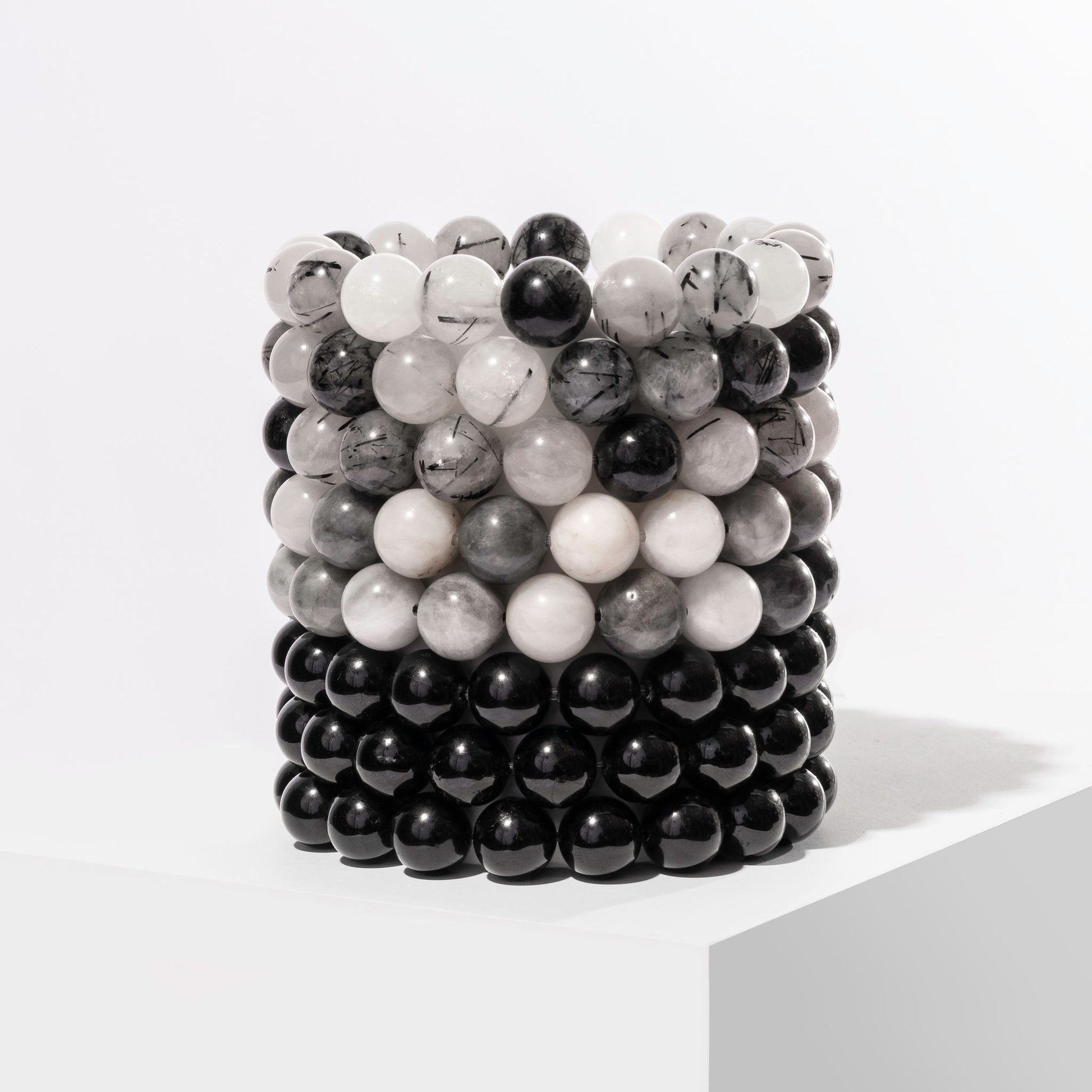 A vertical stack of 8 black and grey crystal gemstone bracelets, positioned on a white block.