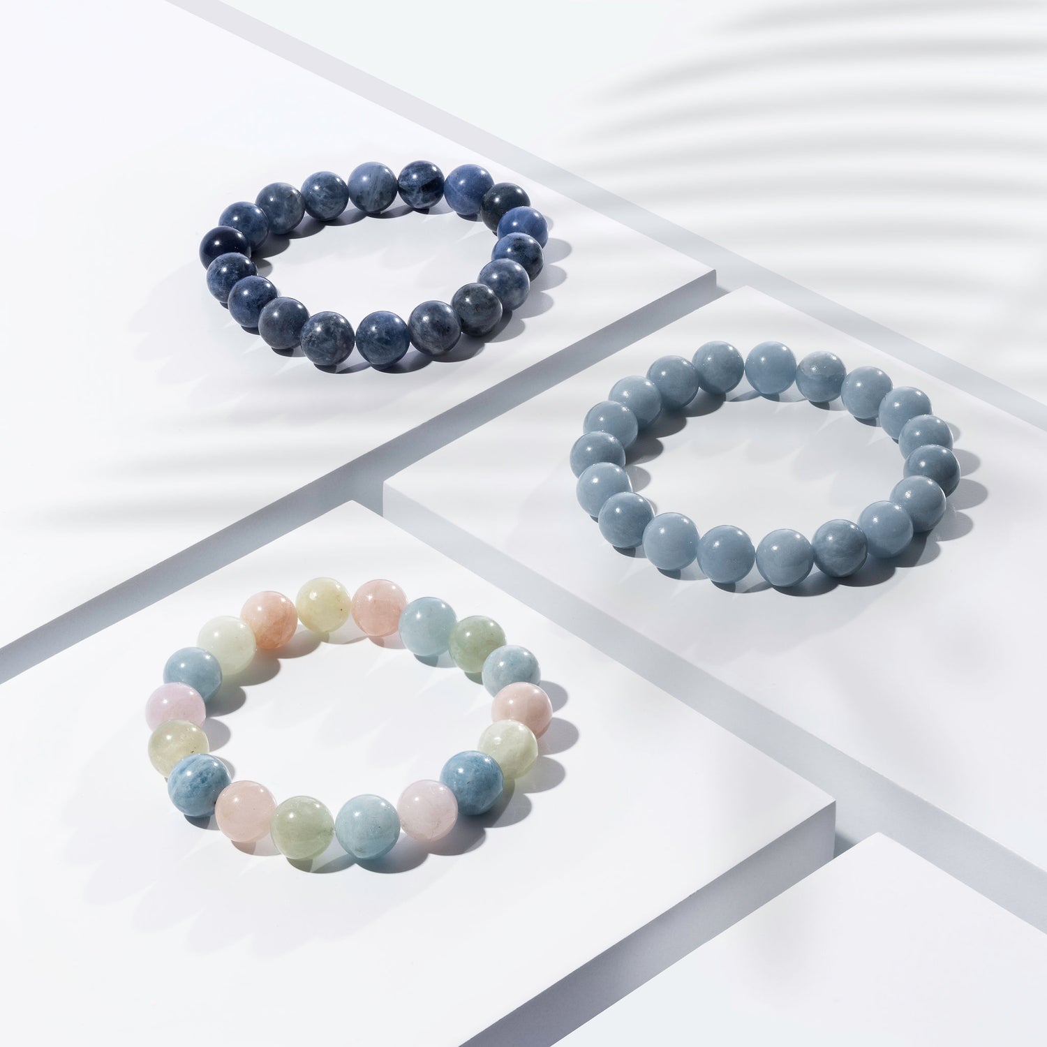 Three blue crystal gemstone bracelets of various shades positioned on white blocks. There is a palm foliage shadow dappling light across the display.