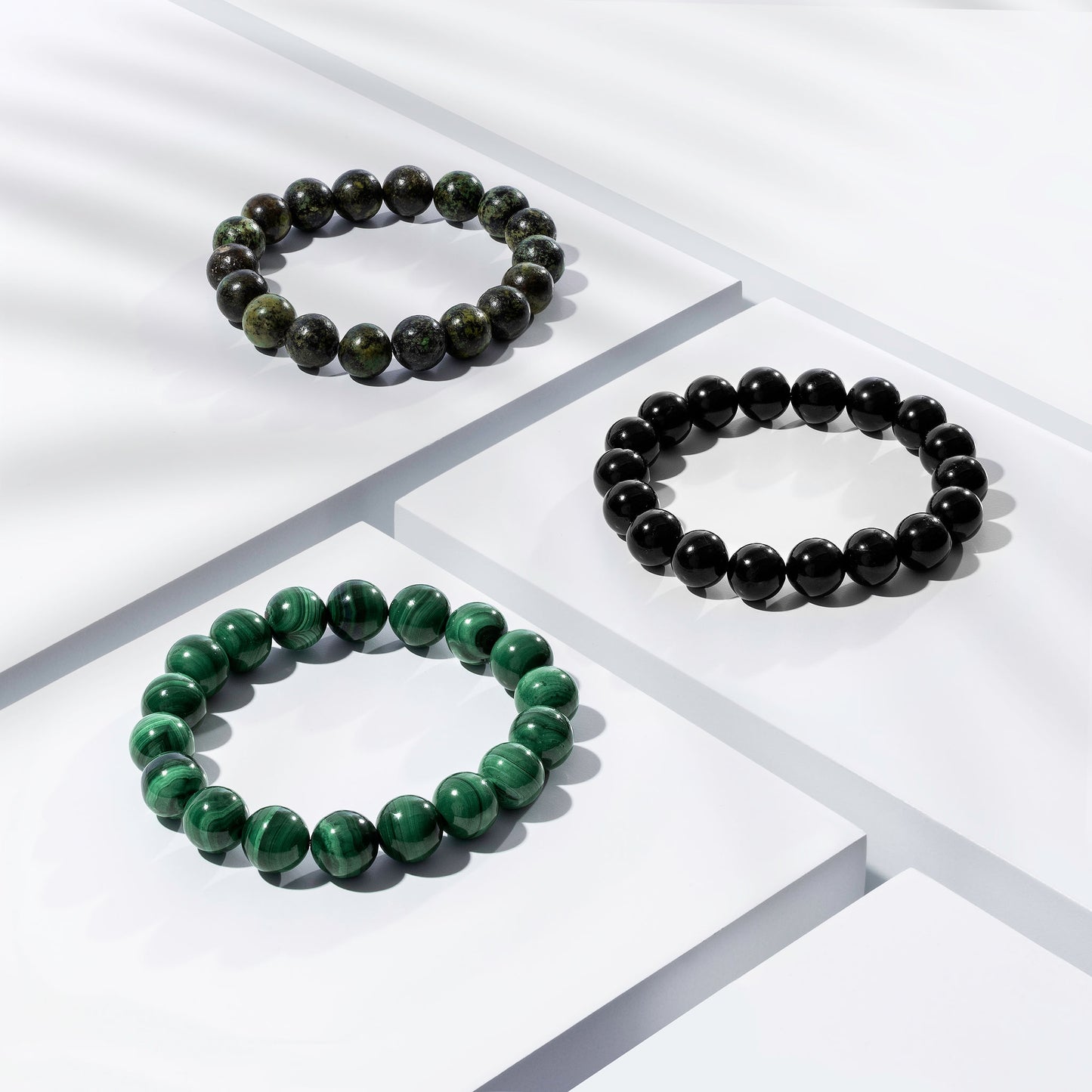 Three coloured crystal gemstone bracelets positioned on white blocks. Two green, and one black. There is a palm foliage shadow dappling light across the display.