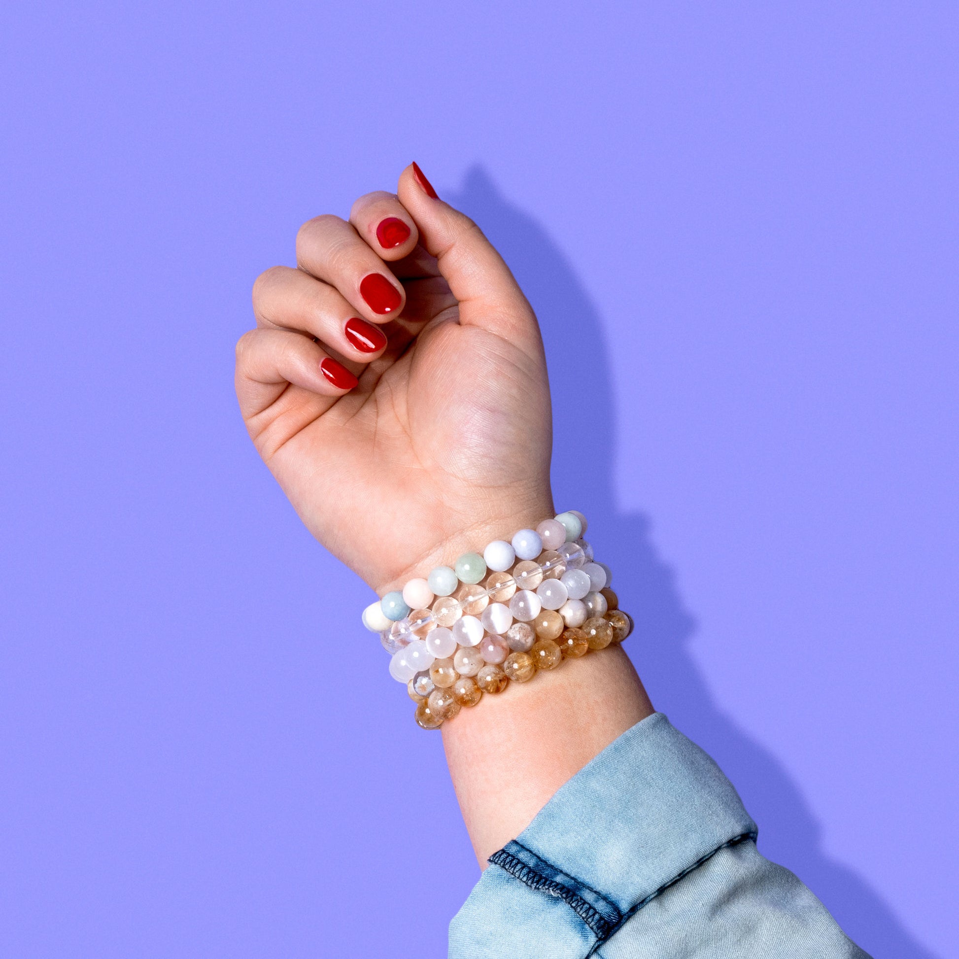 A closeup shot of a stack of yellow, white, and pastel crystal gemstone bracelets on a hand with bright red nail polish.