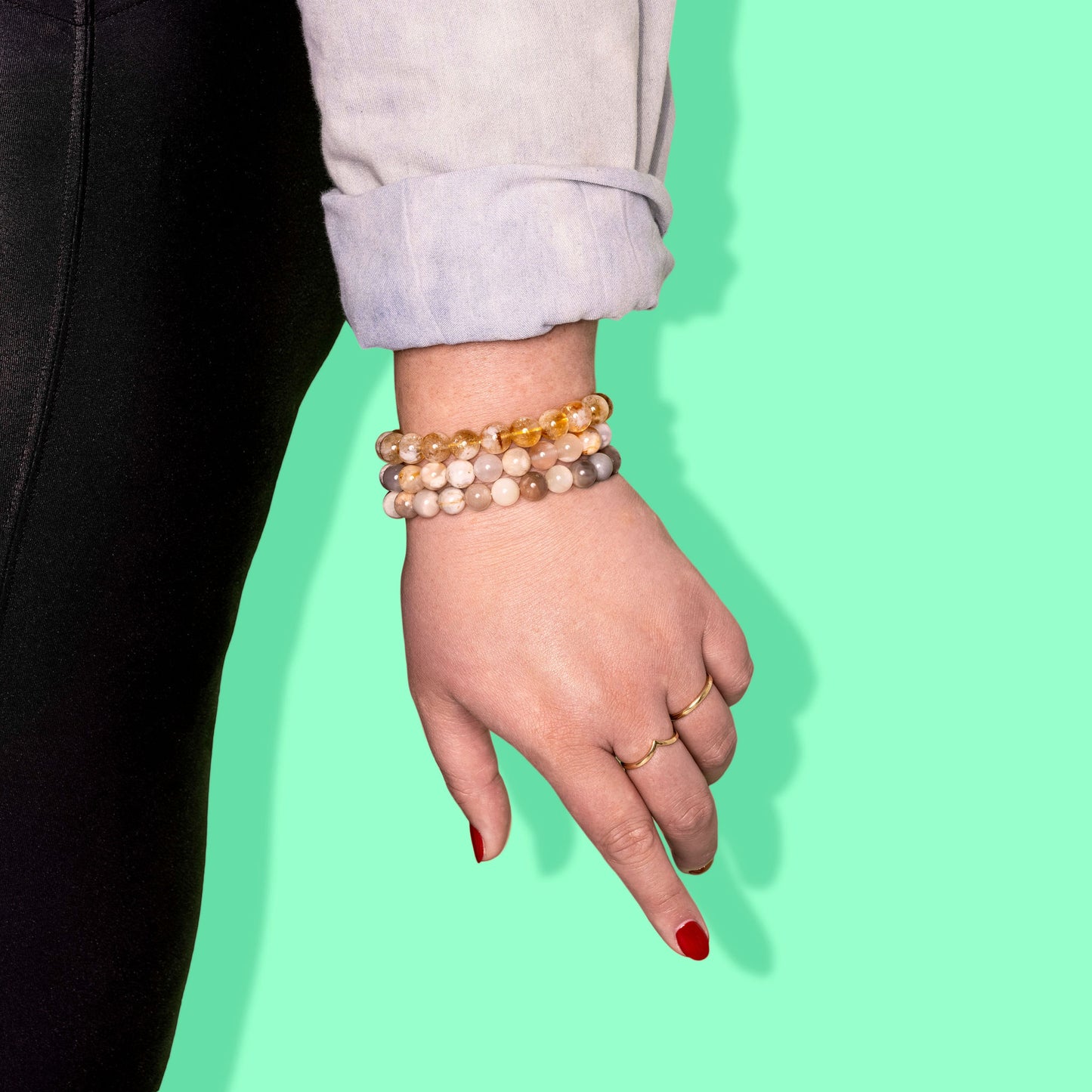 A close-up side profile of a woman wearing black leggings. She wears two gold rings and three yellow and cream coloured crystal gemstone bracelets. The background is bright green.