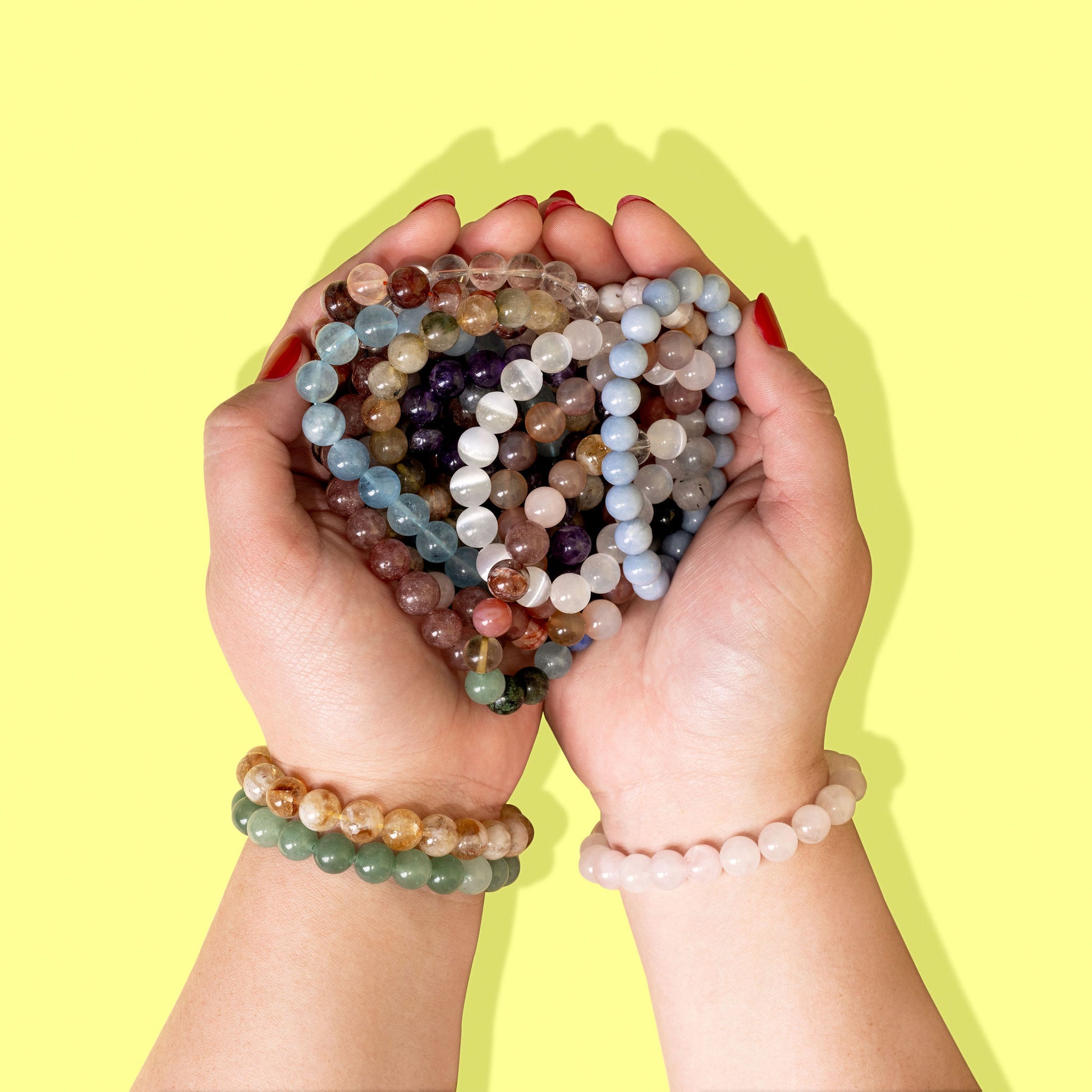 An overhead shot of cupped hands wearing multiple crystal gemstone bracelets. The hands are overflowing with a a pile of brightly coloured crystal gemstone bracelets.