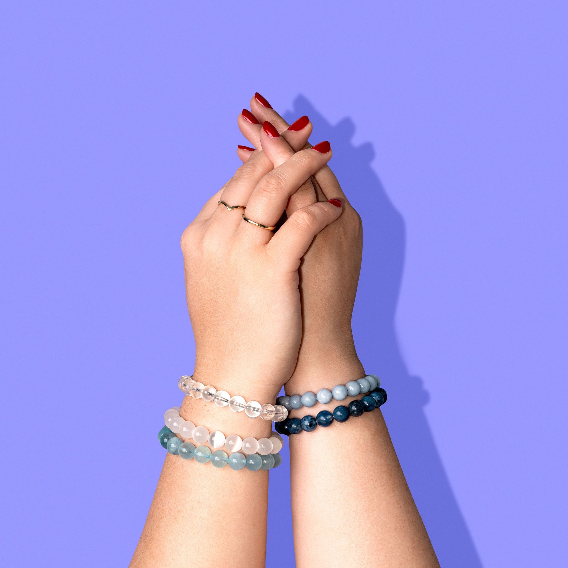 A closeup shot two clasped hands with bright red nail polish wearing crystal and blue gemstone bracelets.