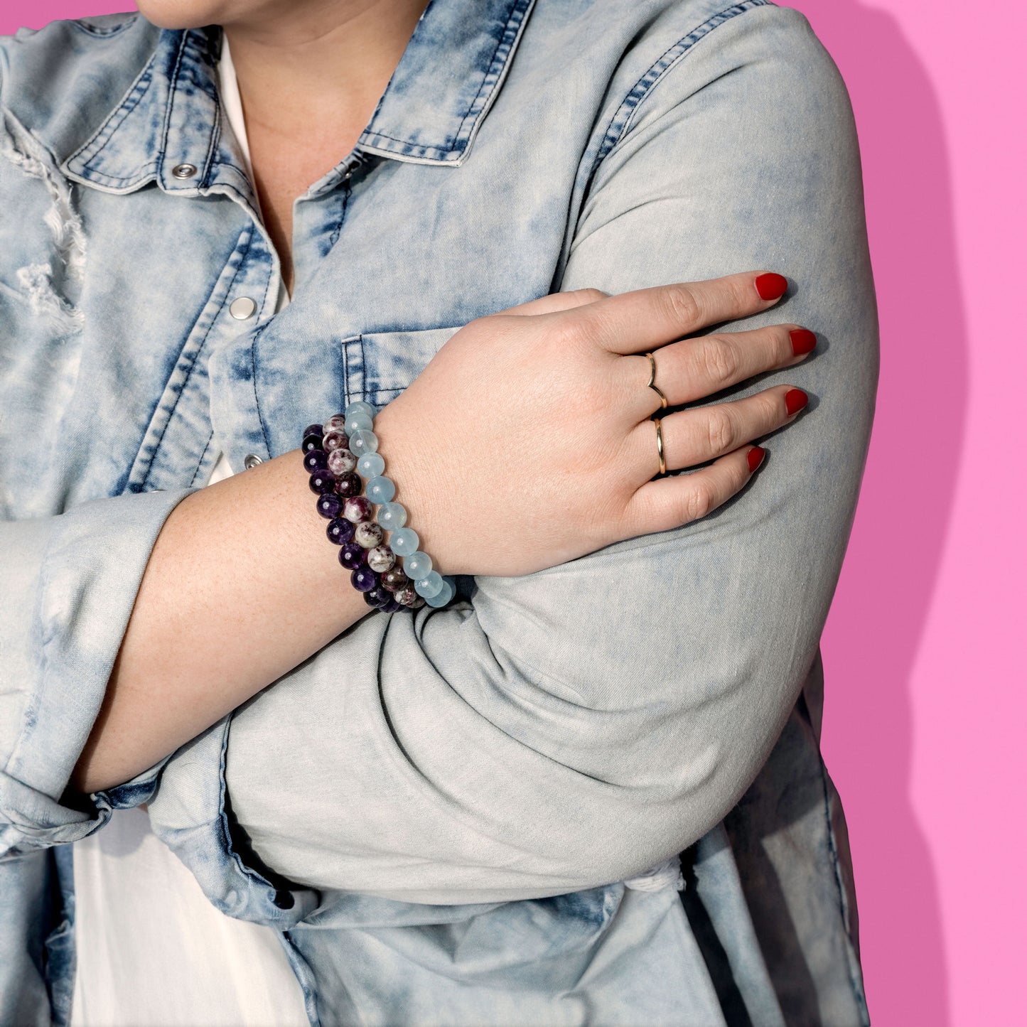 A woman in a distressed denim shirt with bright nail polish and two gold rings wears three blue and purple crystal gemstone bracelets.