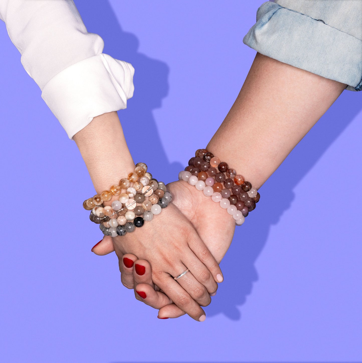 A close up of people holding hands on a bright periwinkle. Each wrist is adorned with multiple crystal gemstone bracelets each in a different colour of red, pink, yellow, and grey.