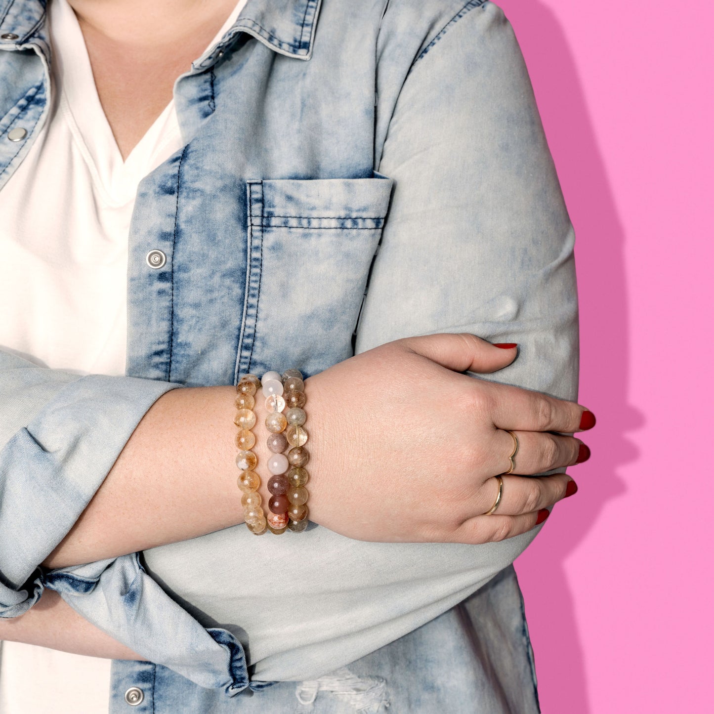 A woman in a white tshirt and distressed denim shirt has her arms crossed. She wears three yellow and cream coloured crystal gemstone bracelets.