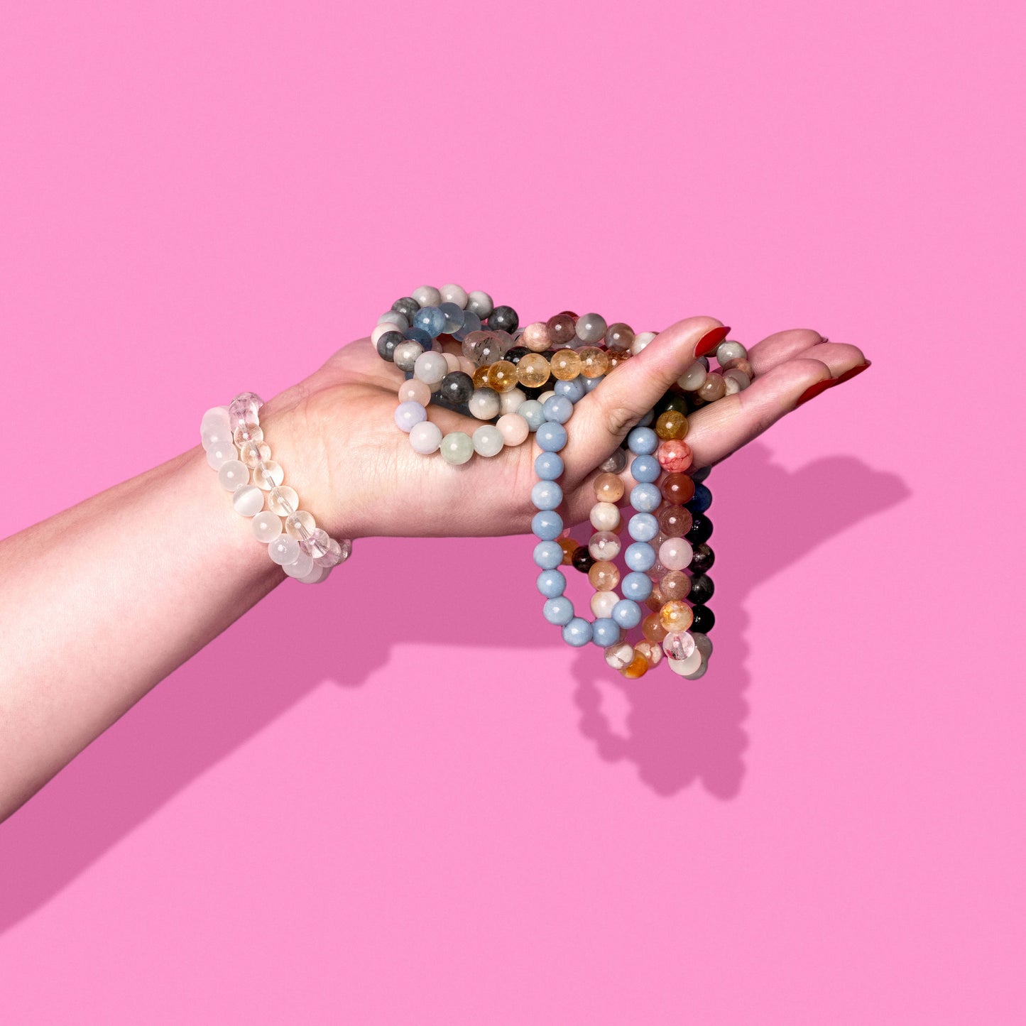 A side profile of a hand on a bright pink background. It wears several crystal bracelets and holds an overflowing pile of colourful gemstone bracelets.