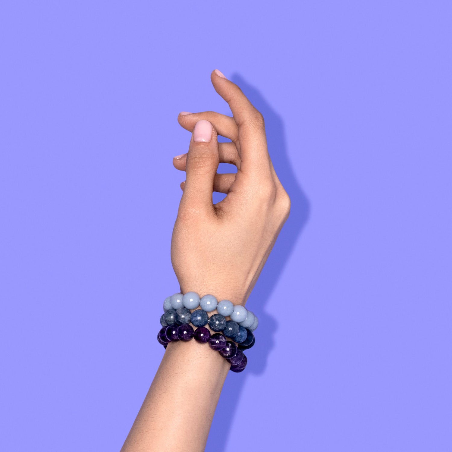 A hand with light pink nail polish wearing 3 different crystal gemstone bracelets blue and purple in colour. The background is bright periwinkle with a hard shadow to the right.