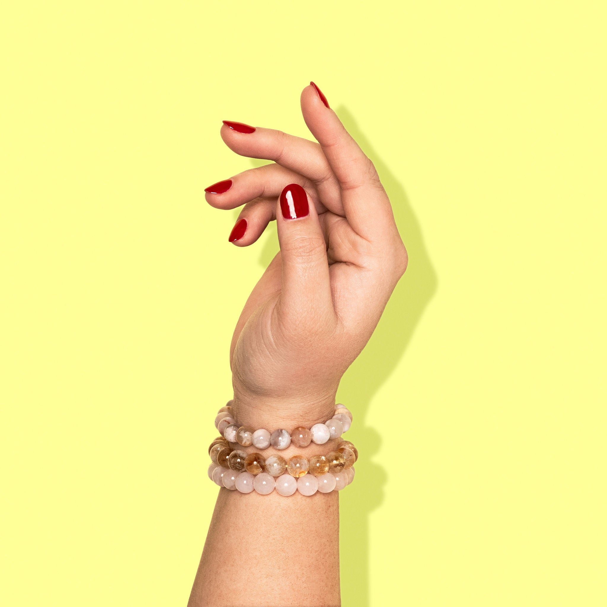 A closeup shot of a stack of yellow, white, and pink crystal gemstone bracelets on a hand with bright red nail polish.