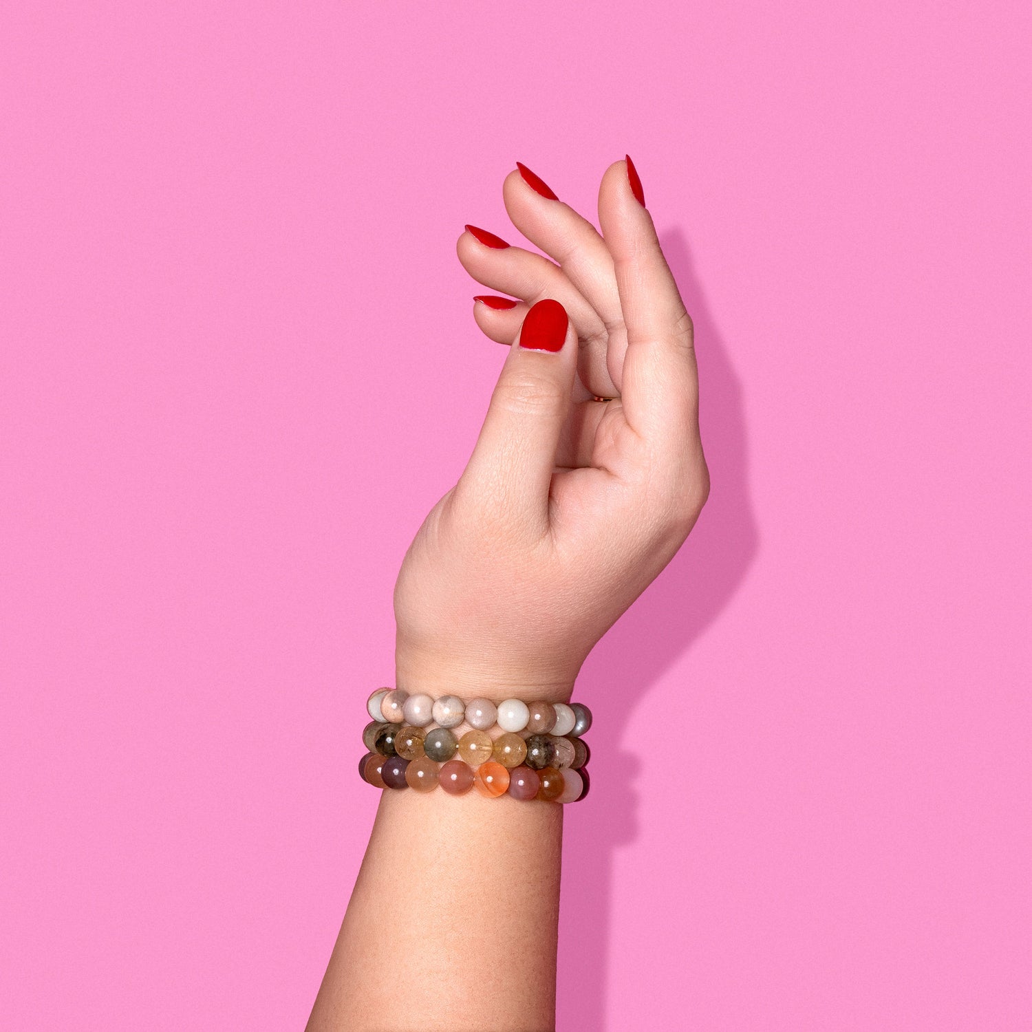 A hand with bright red nails wearing 3 different pink, yellow, and white crystal gemstone bracelets. The background is bright pink with a hard shadow to the right.