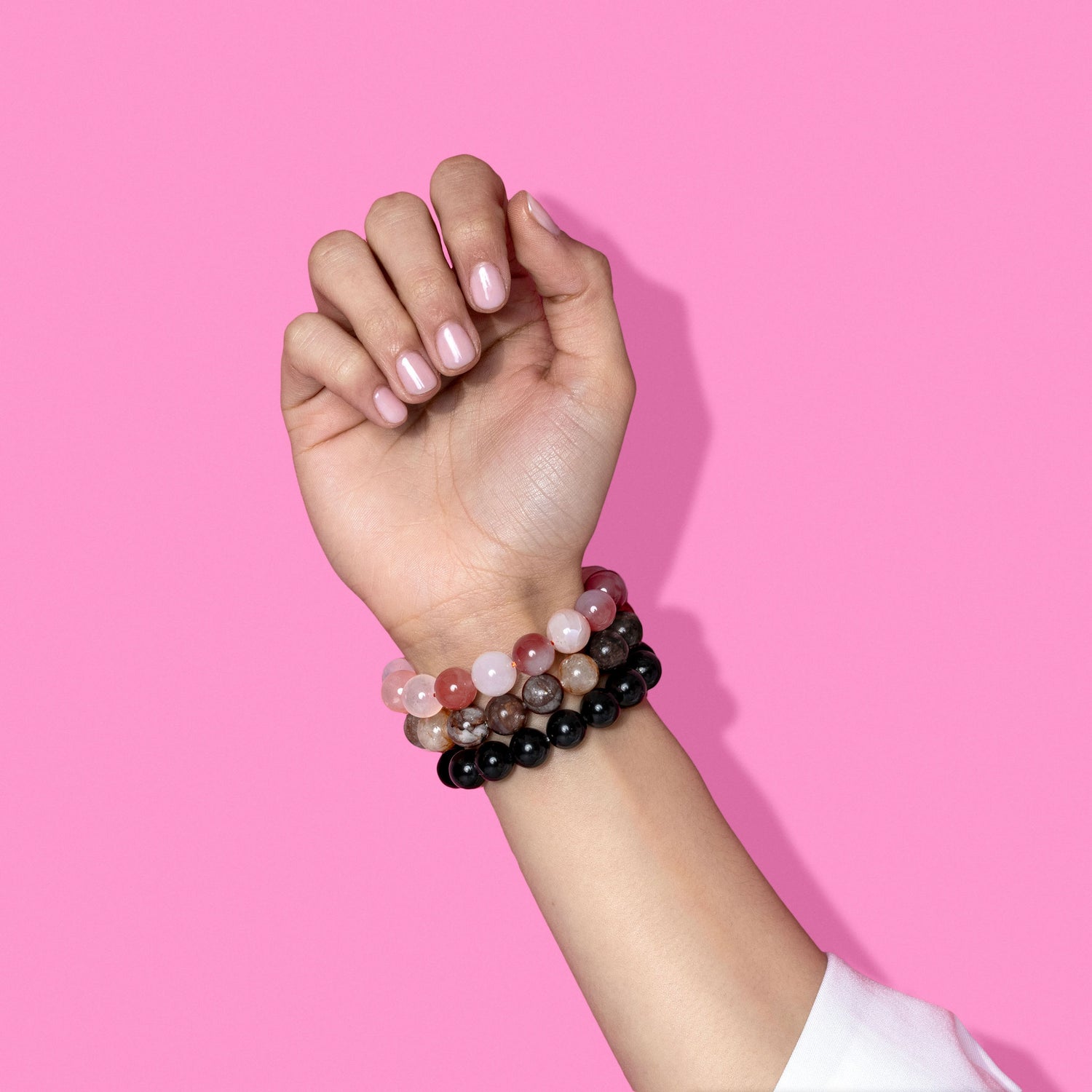 A closeup shot of a stack of pink, purple, and black crystal gemstone bracelets on a hand with light pink nail polish.