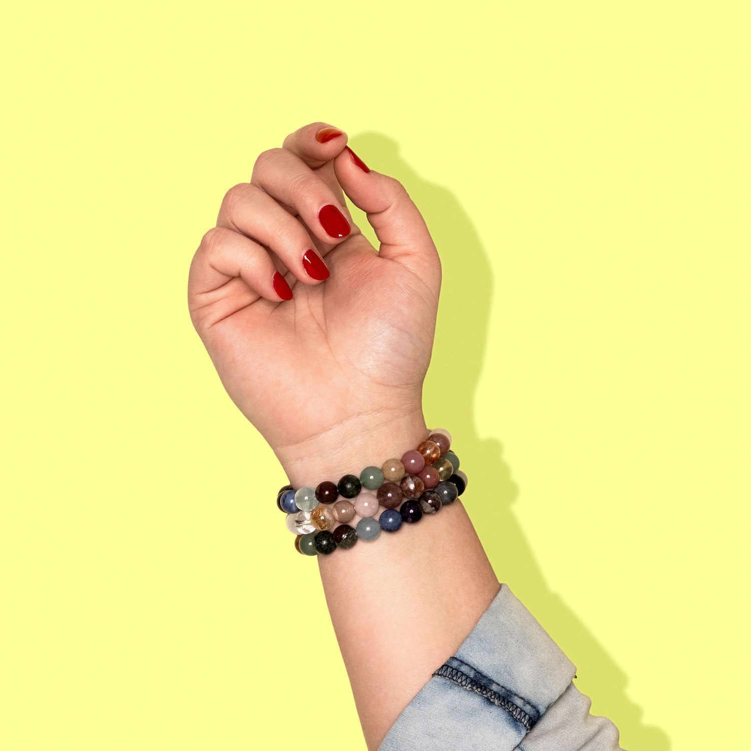 A closeup shot of a stack of multicoloured crystal gemstone bracelets on a hand with bright red nail polish.