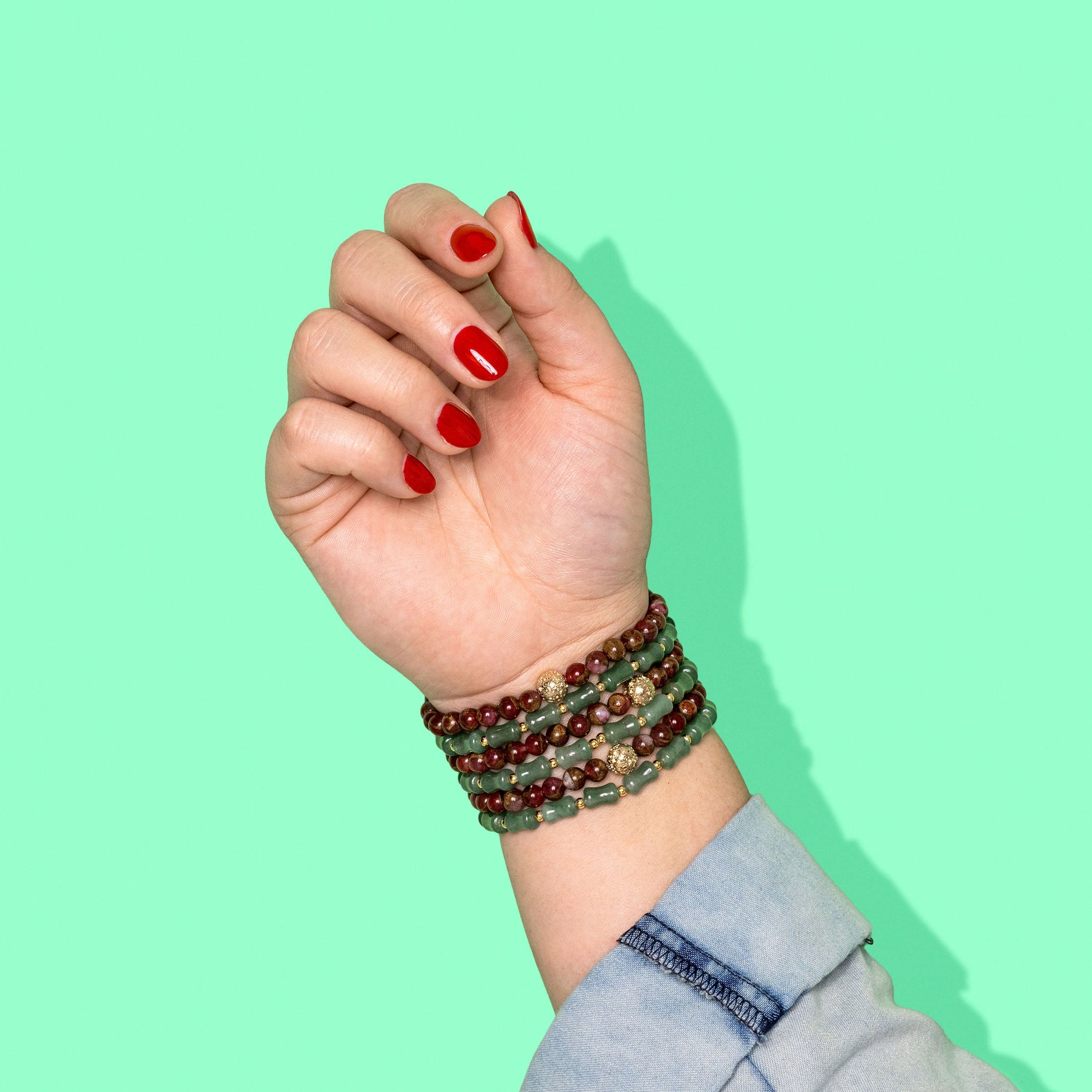 A closeup shot of a stack of green bamboo shaped beads and red copper agate crystal gemstone bead bracelets on a hand with bright red nail polish.